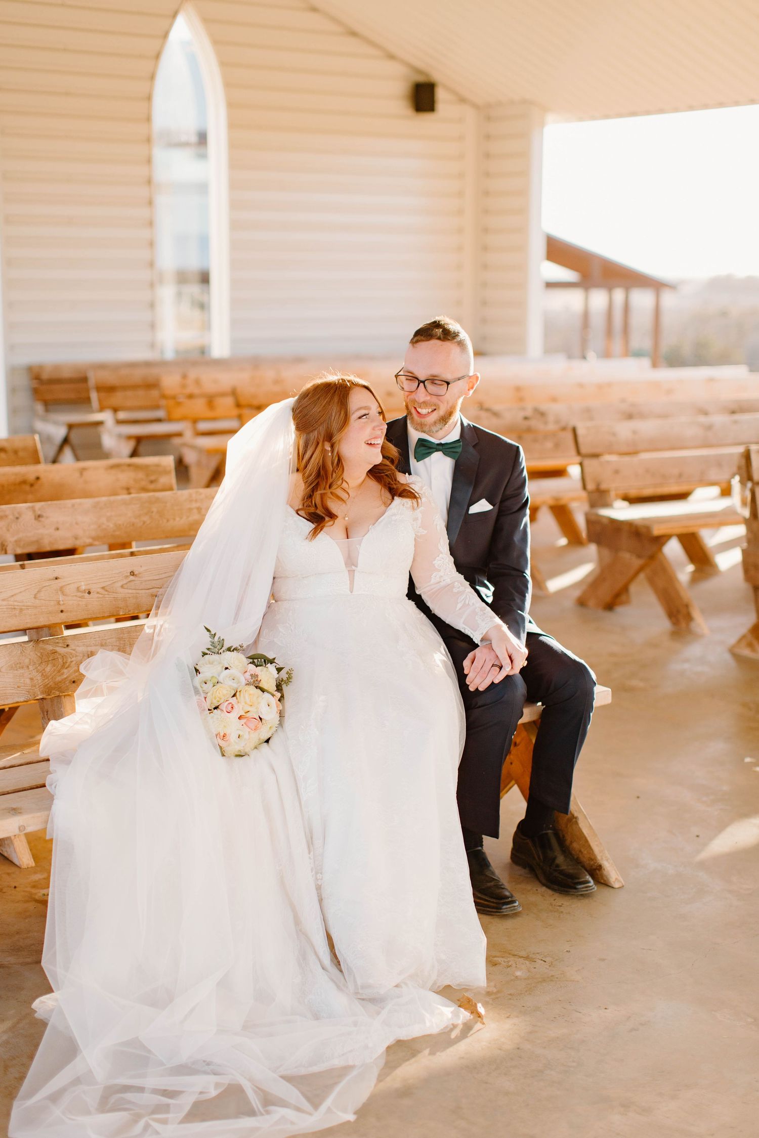 Bride and groom sitting together on wooden pews outside a white chapel wedding venue.