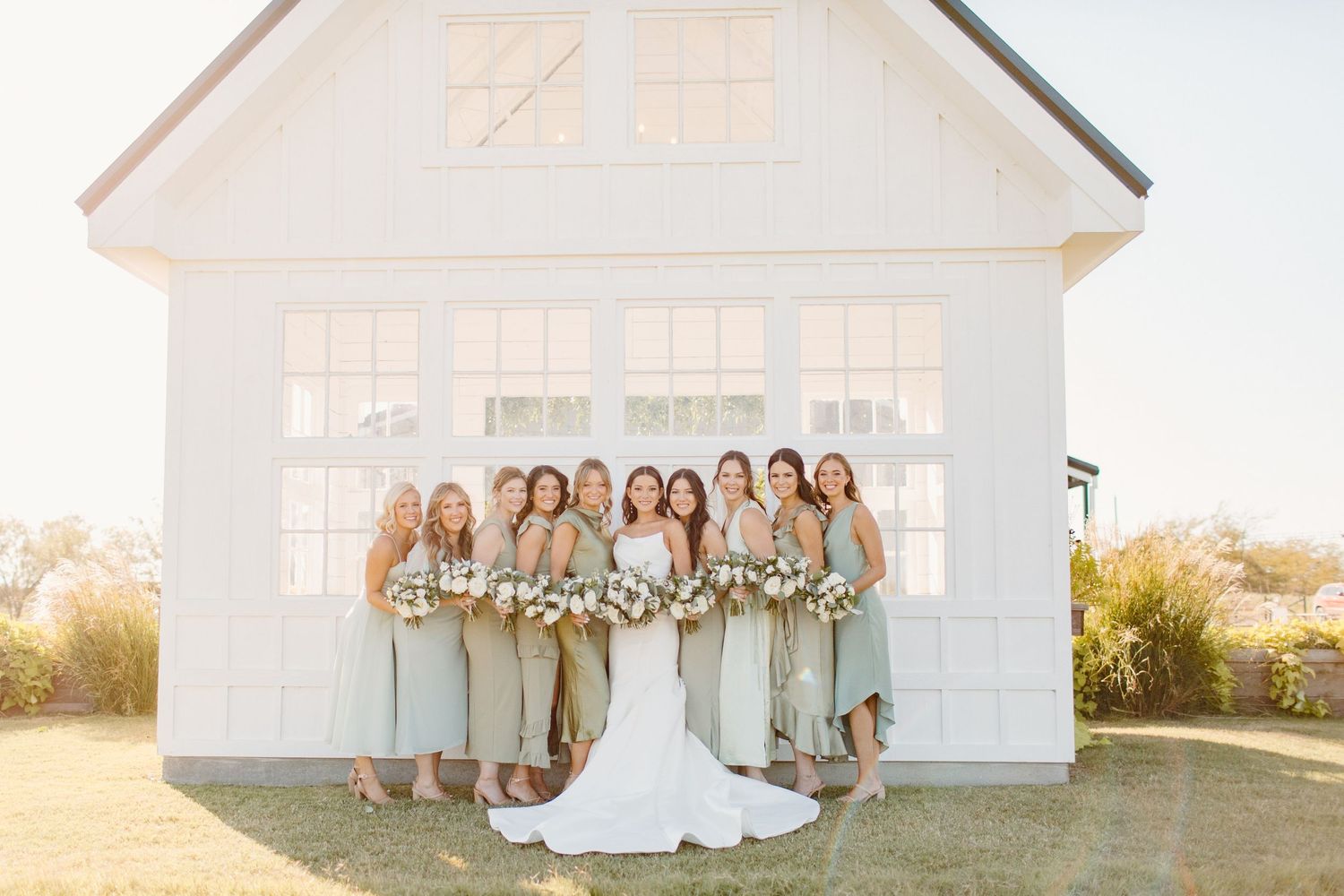 Bride in white gown stands with eight bridesmaids in sage green dresses in front of a white barn venue.