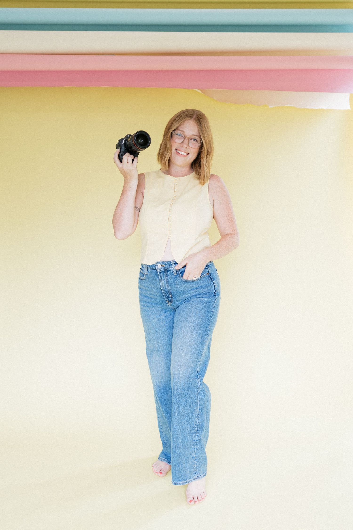 A smiling female photographer holding a camera in a yellow studio with colorful striped backdrop.