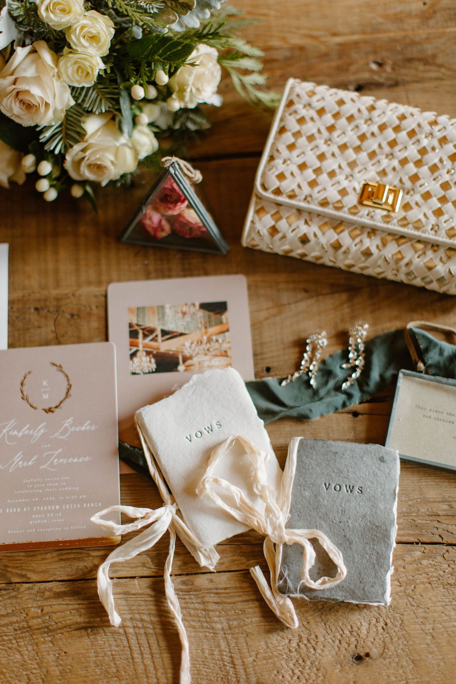 Wedding flat lay featuring vow books, floral invitation suite, bridal clutch, and white roses.