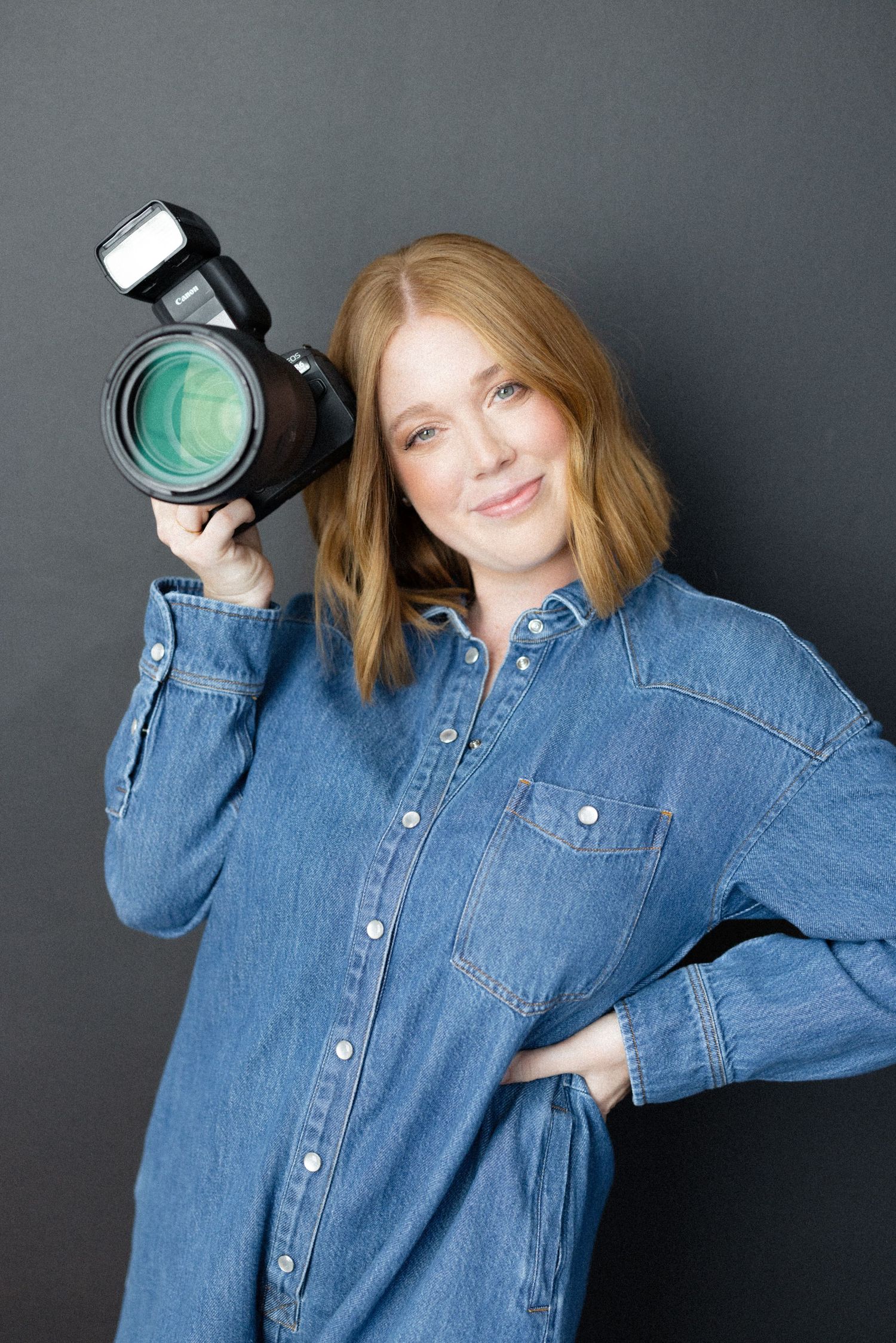 A smiling female photographer in a denim shirt holds a DSLR camera with flash against a gray background.