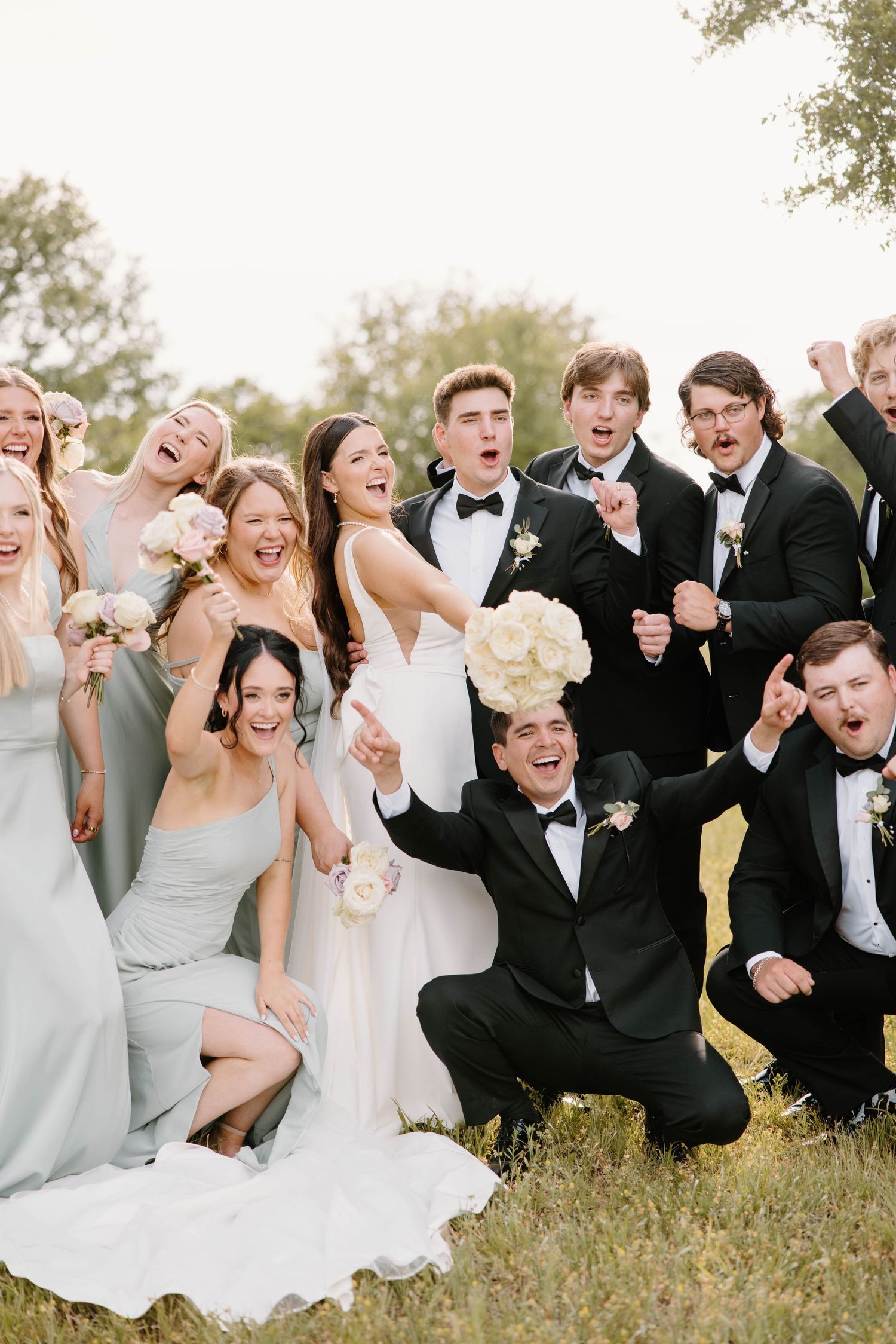 A joyful wedding party in sage green dresses and black tuxedos celebrates together on a lawn.