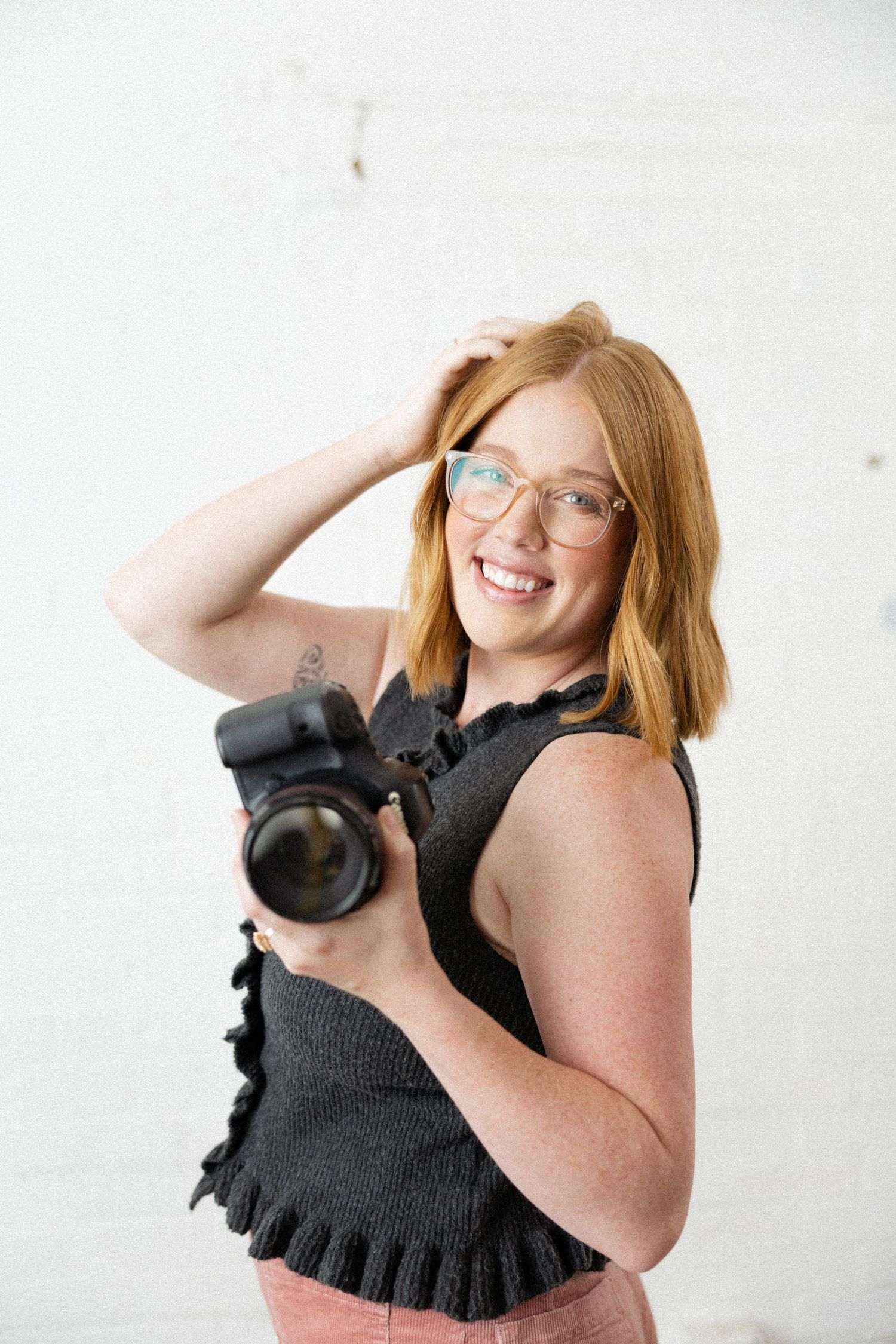 Smiling redhead female photographer holding a professional DSLR camera against a white brick wall background.