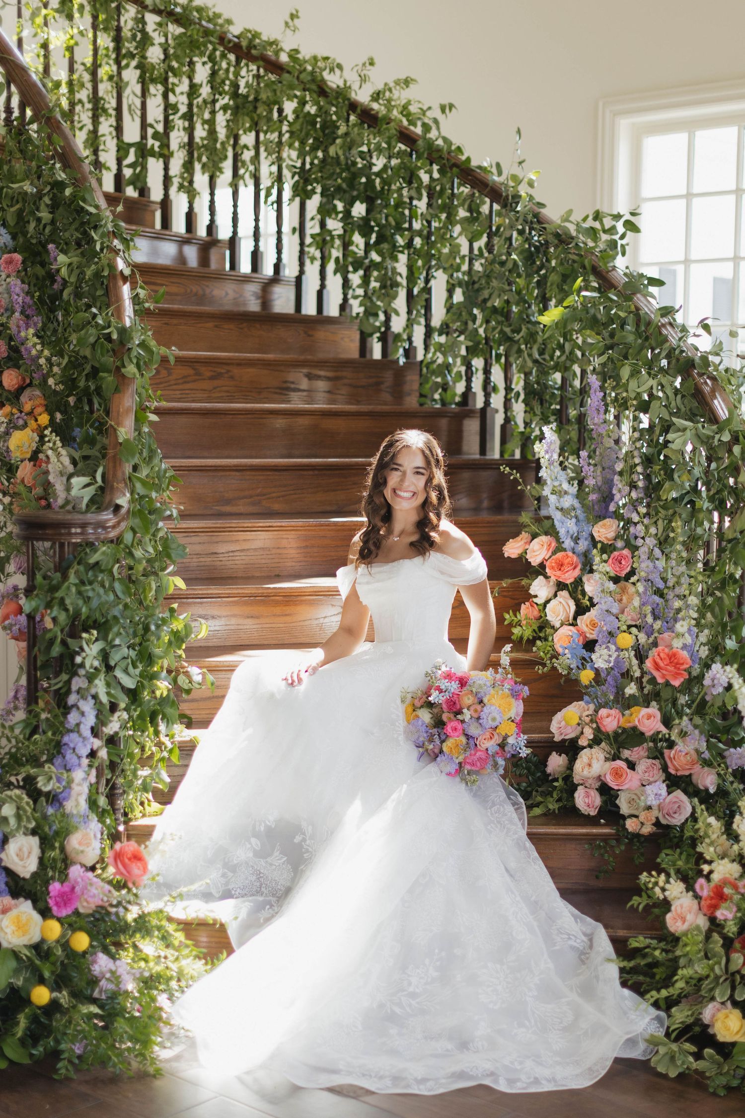 Bride in white off-shoulder gown sits on floral-decorated staircase holding colorful bouquet.