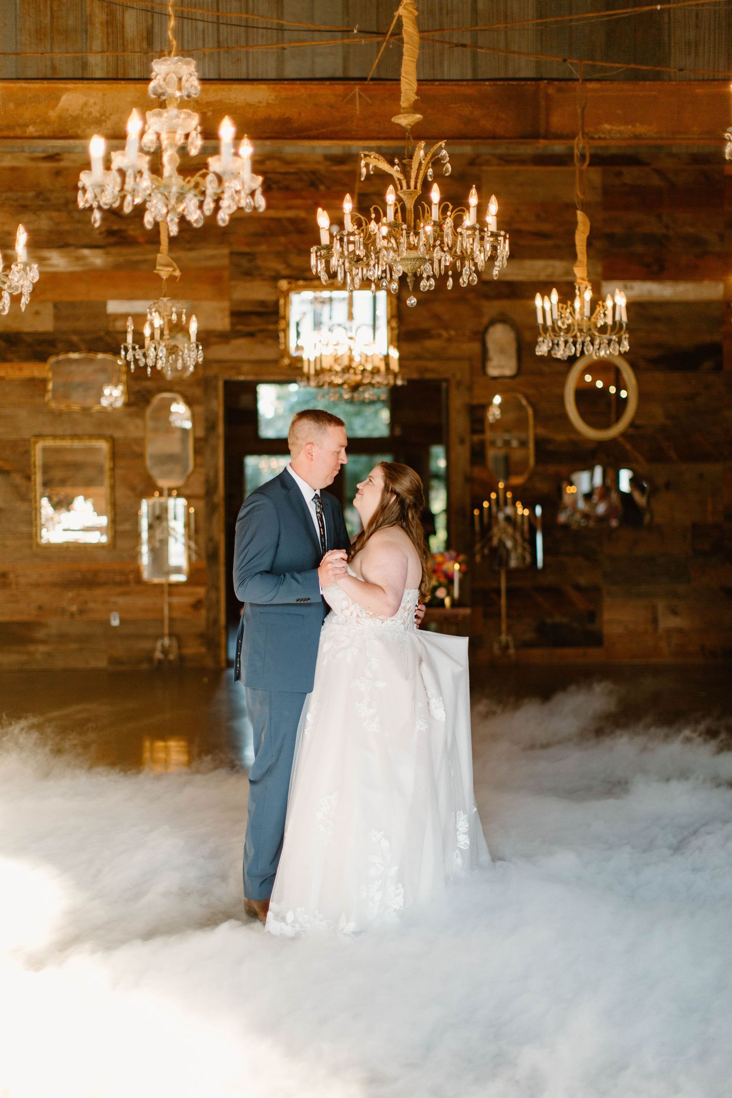 Bride and groom share first dance surrounded by dramatic fog under sparkling chandeliers in a rustic barn venue.