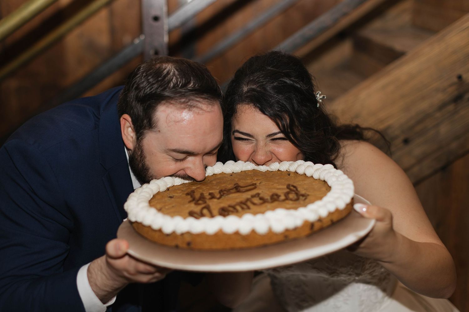 Bride and groom playfully bite into a round cookie cake inscribed with Just Married at their wedding reception.