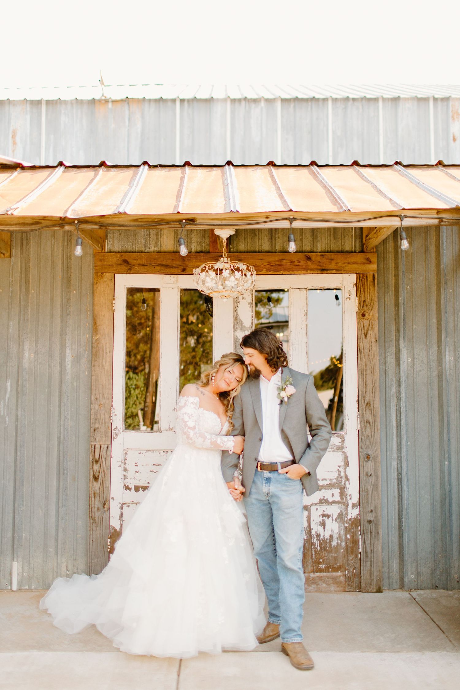 Bride in lacy ballgown and groom in gray suit with jeans share a romantic moment outside a rustic barn venue.
