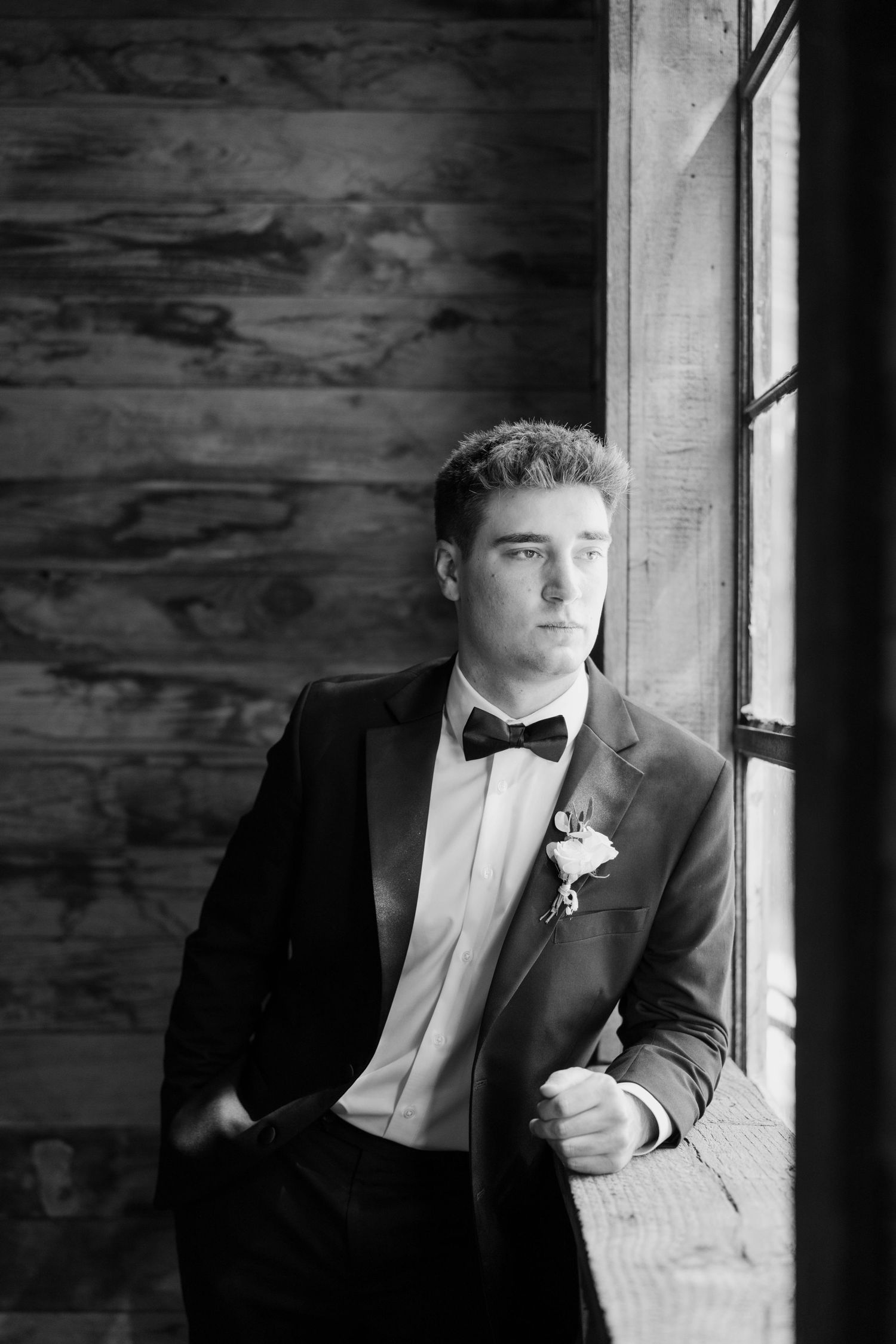 Black and white portrait of groom in tuxedo leaning against window in rustic barn.