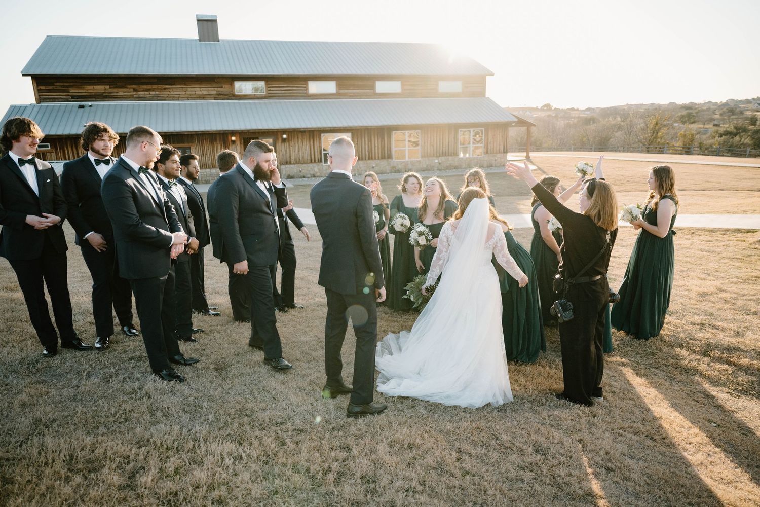 A wedding party gathers in golden hour light outside a rustic barn venue for a group celebration.