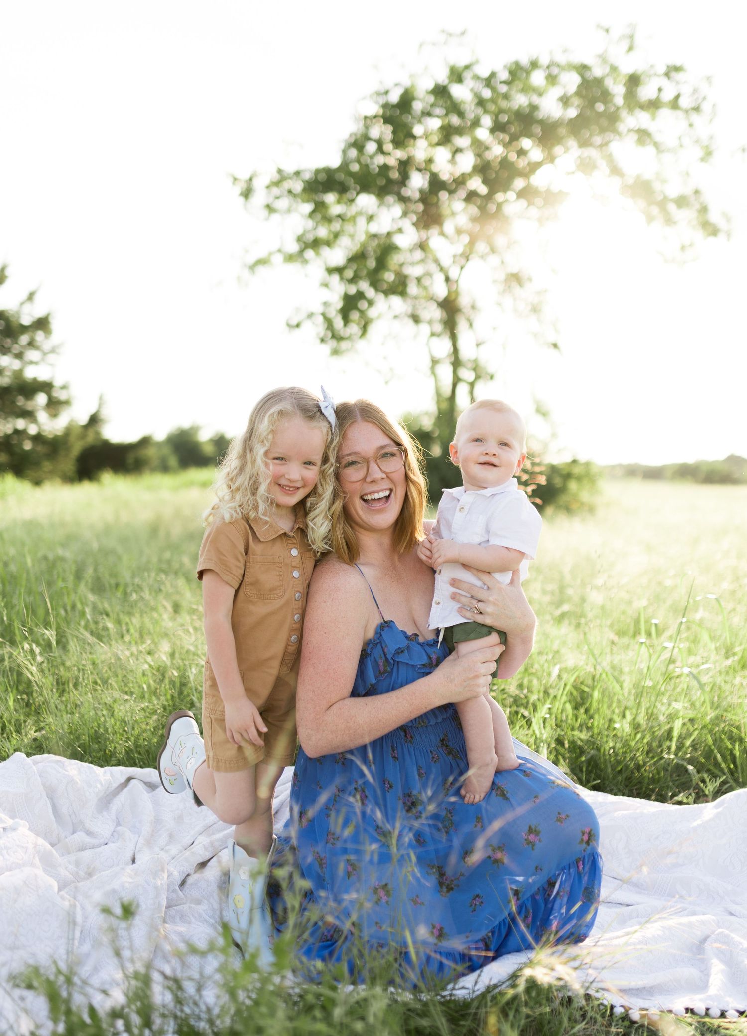 A joyful mother sits in a sunlit green field with her two young children, wearing a blue floral dress.