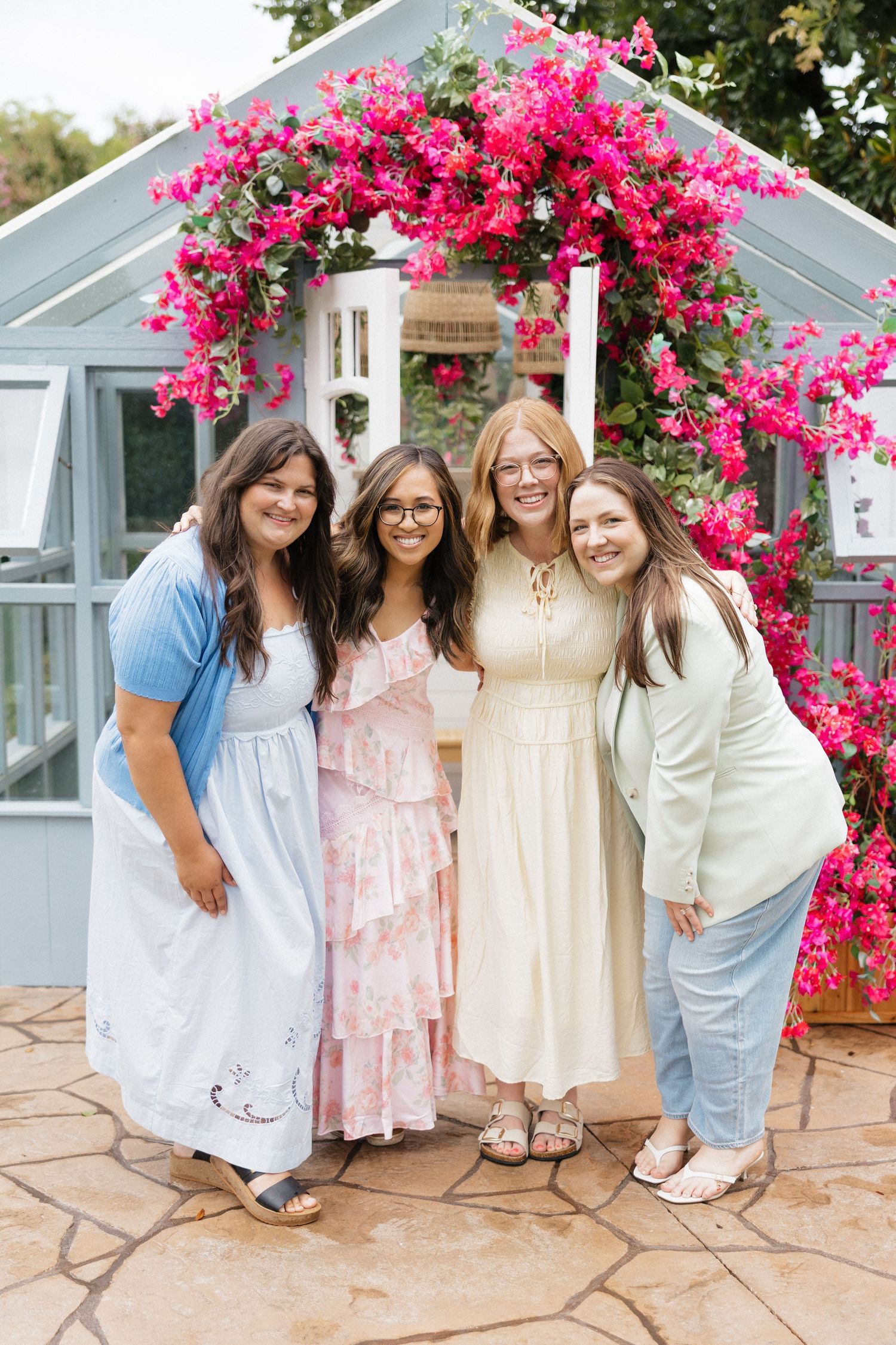 Four women in elegant dresses posing together under a vibrant pink bougainvillea arch outside a greenhouse.