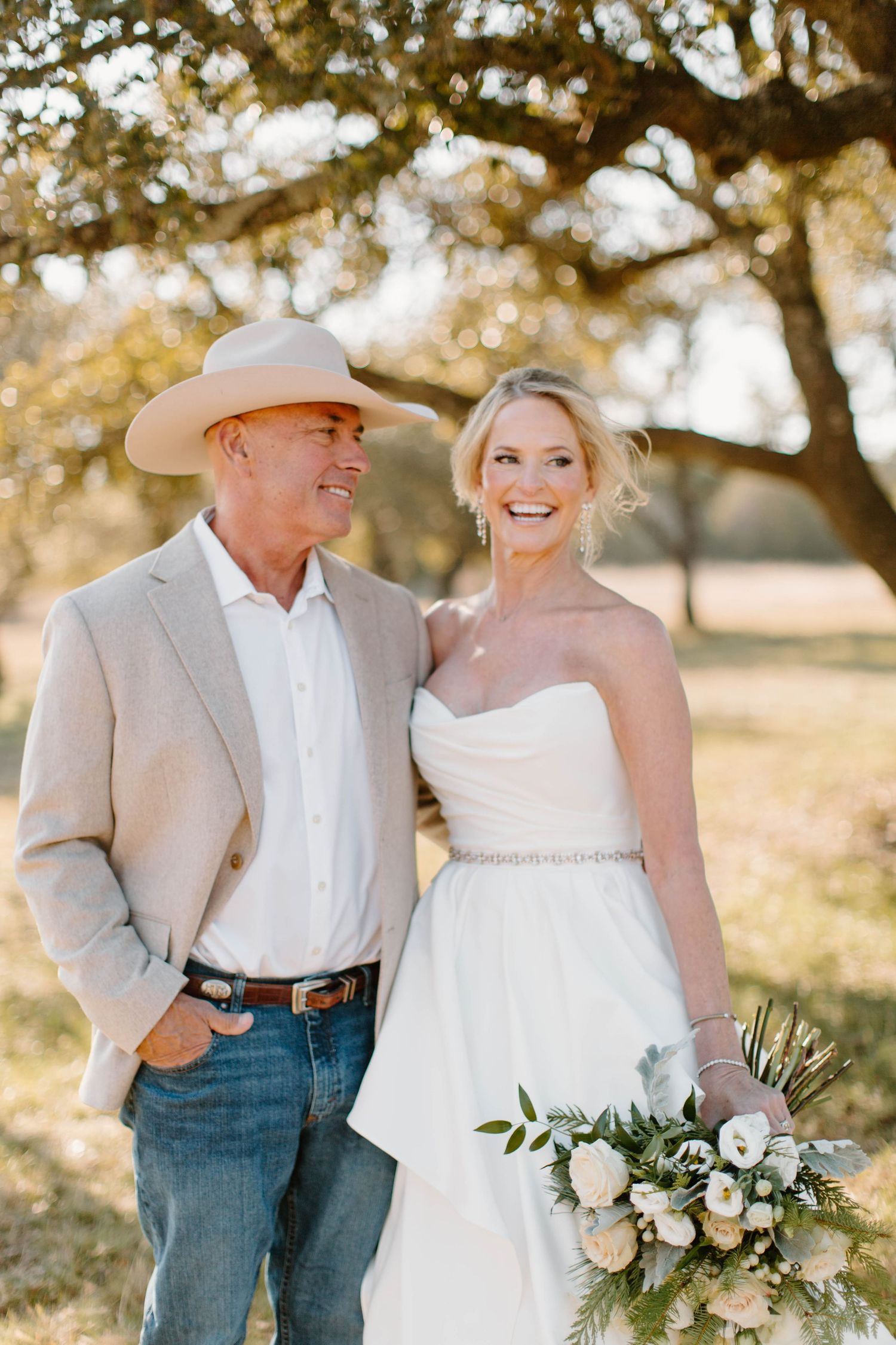 Smiling bride in strapless gown with groom in cowboy hat under oak trees outdoors.