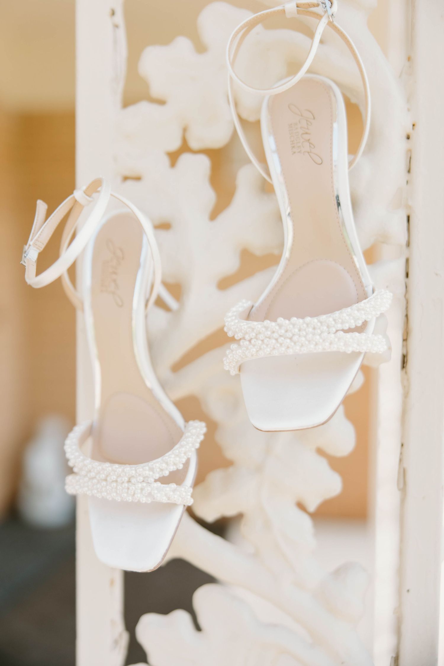 Elegant white pearl-embellished bridal heels hang on a decorative white carved wooden frame.