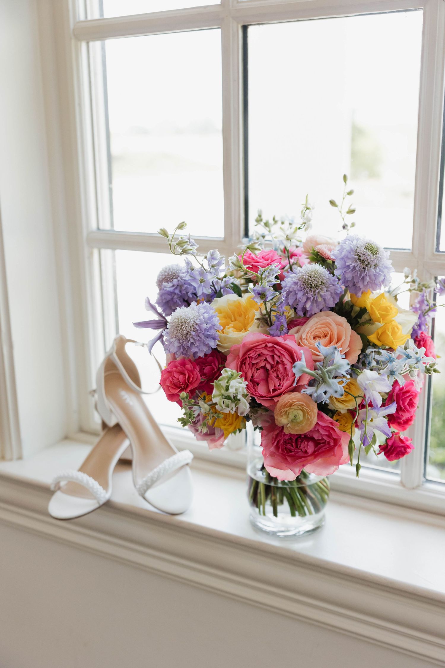 Colorful wedding bouquet with pink, purple, and yellow flowers beside white heels on windowsill.