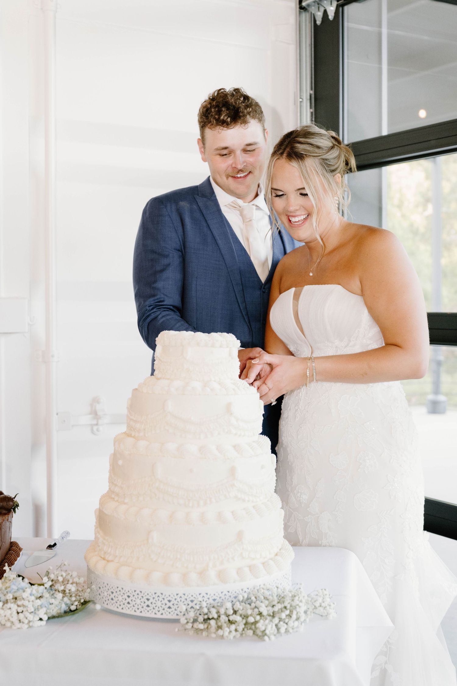Smiling bride and groom cutting four-tier white wedding cake at reception.