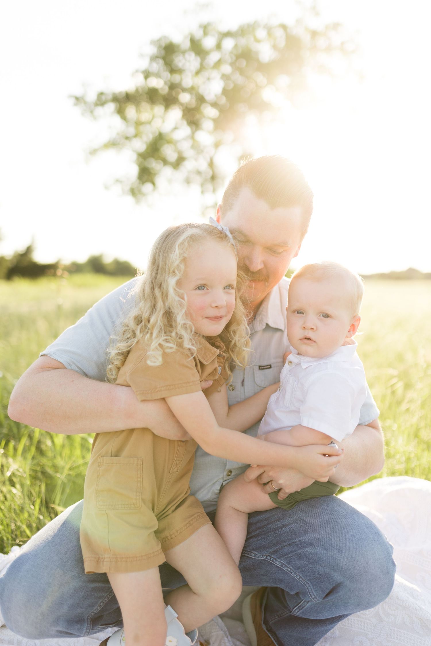 A loving father kneels in a sunny green meadow embracing his young daughter and baby son.