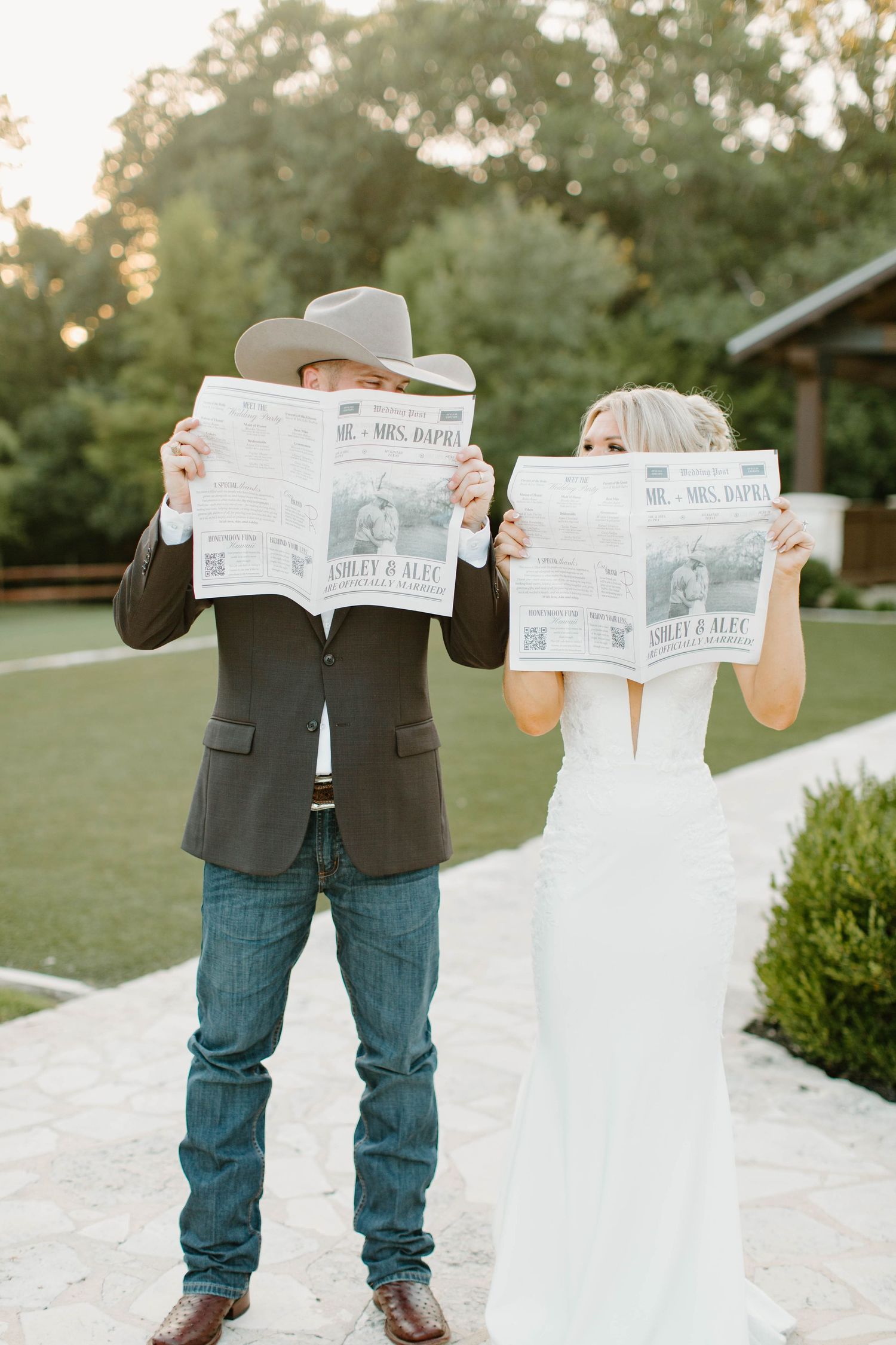 A bride and groom hold personalized wedding newspapers in front of their faces outdoors.