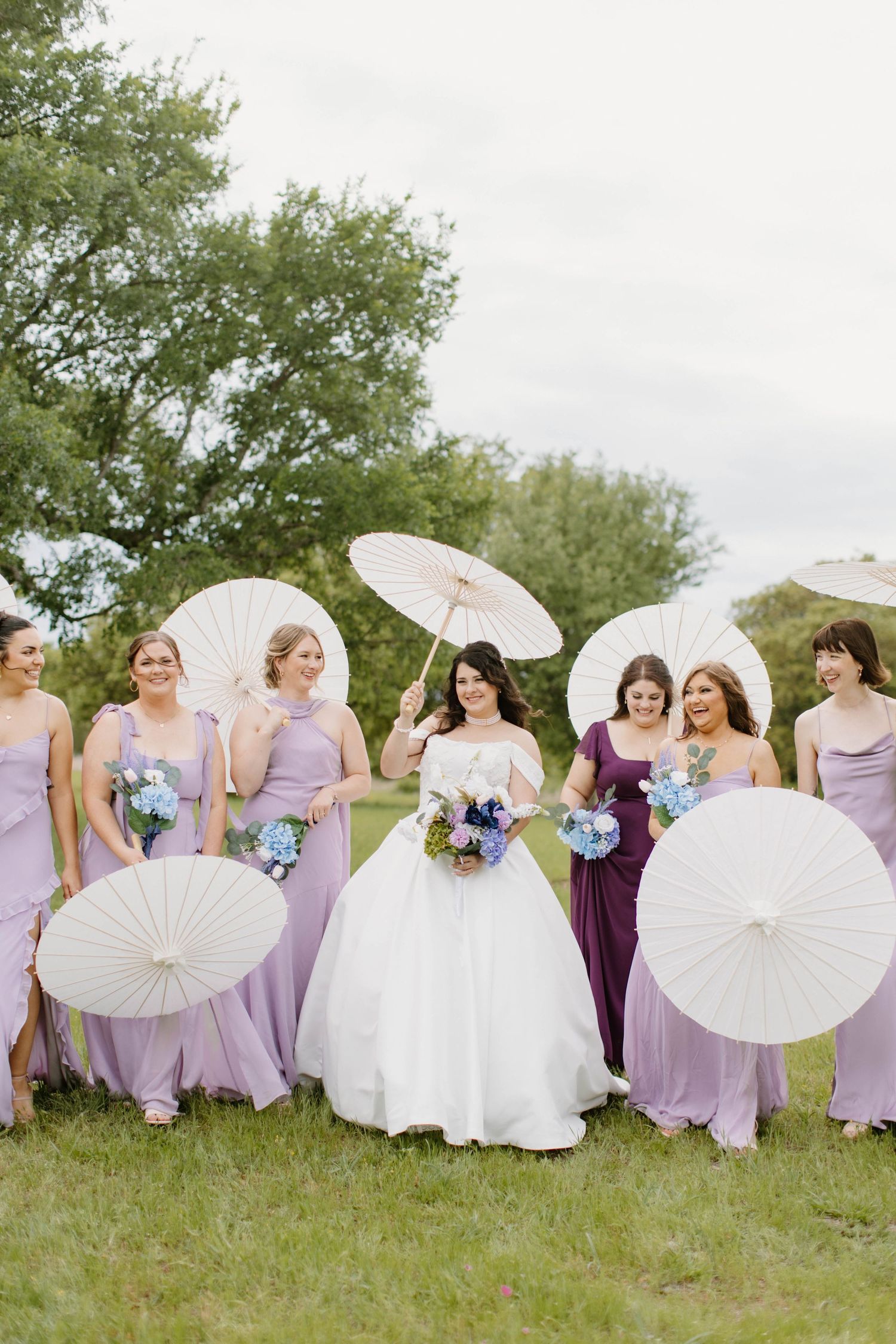 A bride and six bridesmaids in lavender dresses holding white parasols outdoors on a cloudy day.
