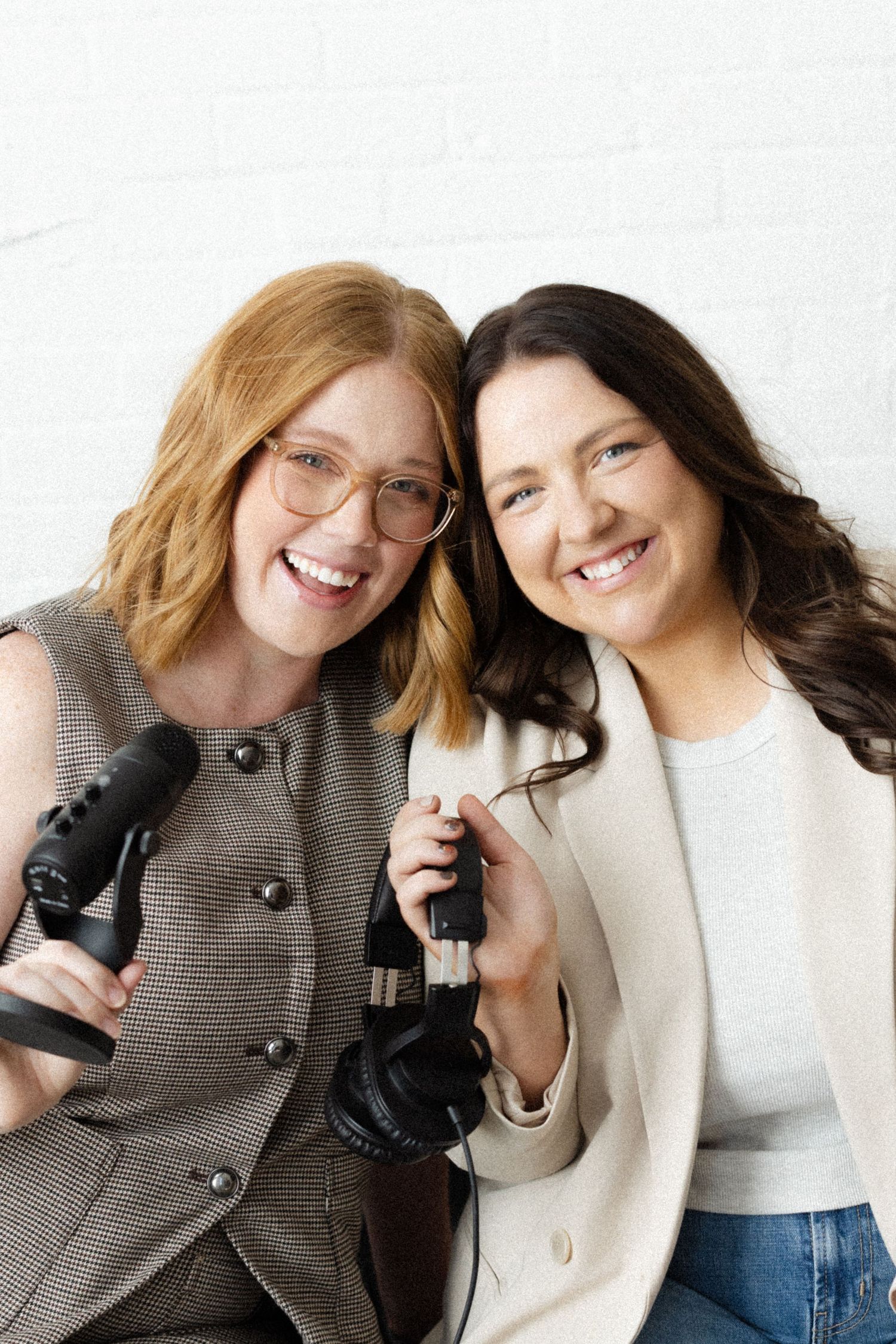 Two smiling women hold podcast microphones while posing together against a white brick wall.