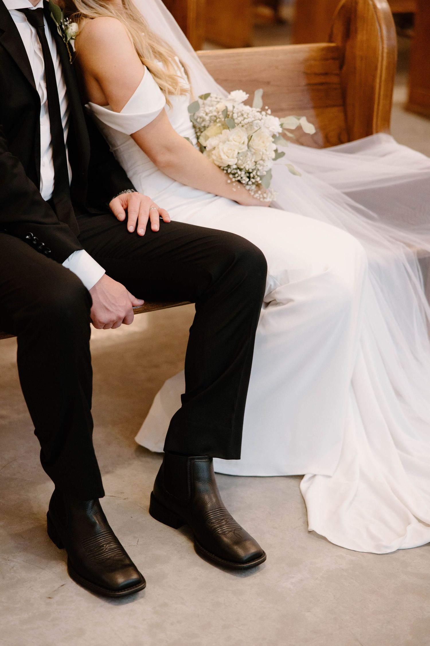 Bride and groom sitting together in church, groom wearing black suit with cowboy boots, bride in white off-shoulder gown holding bouquet.