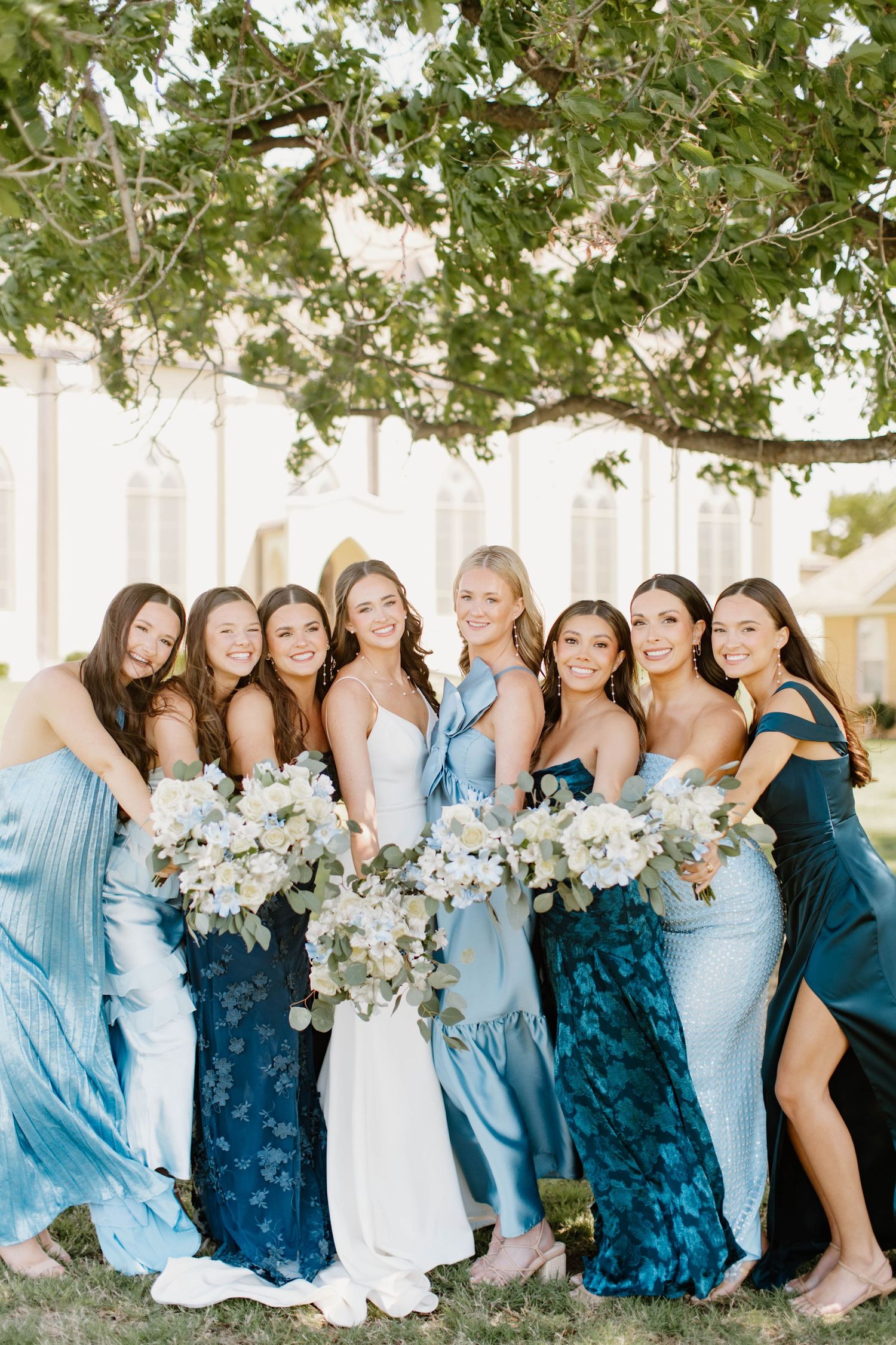 A radiant bride poses with seven bridesmaids in shades of blue dresses holding white floral bouquets.