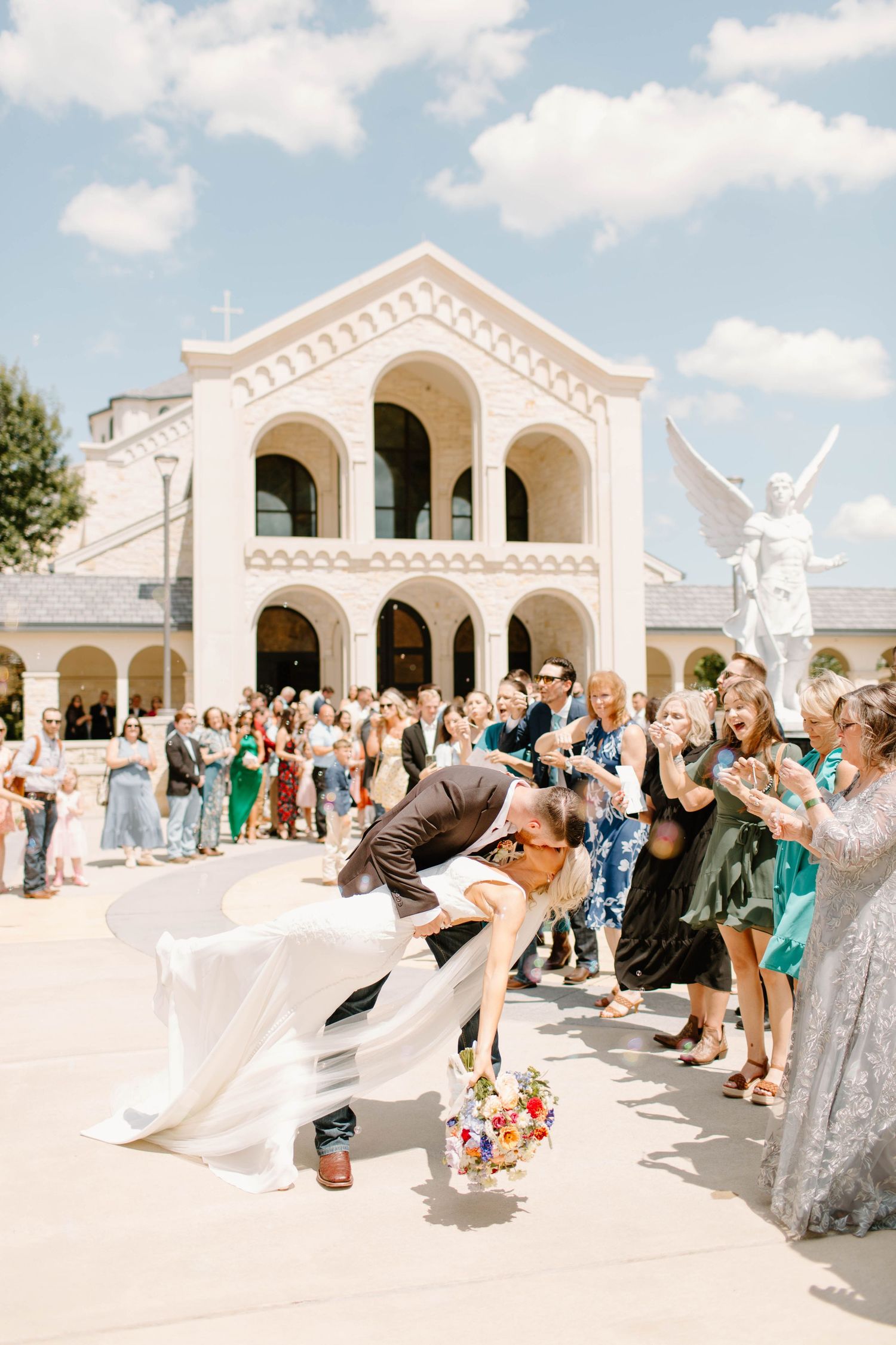A groom dips his bride for a kiss outside a white church while guests cheer around them.