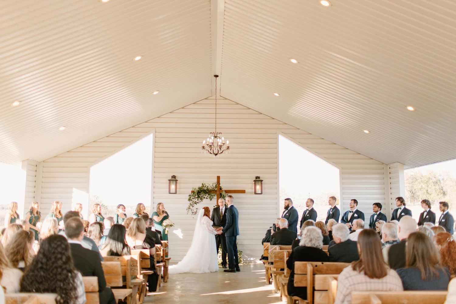 Wedding ceremony inside white chapel with vaulted ceiling, guests seated in wooden pews.