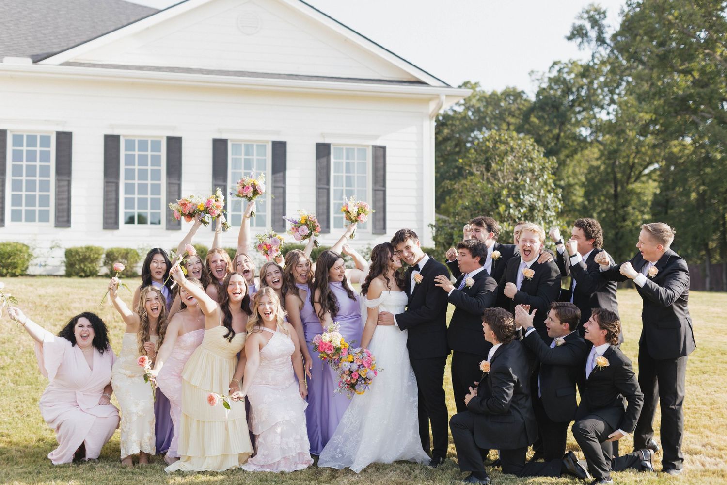 Large wedding party celebrating outdoors in front of a white venue, bridesmaids in purple and groomsmen in black.