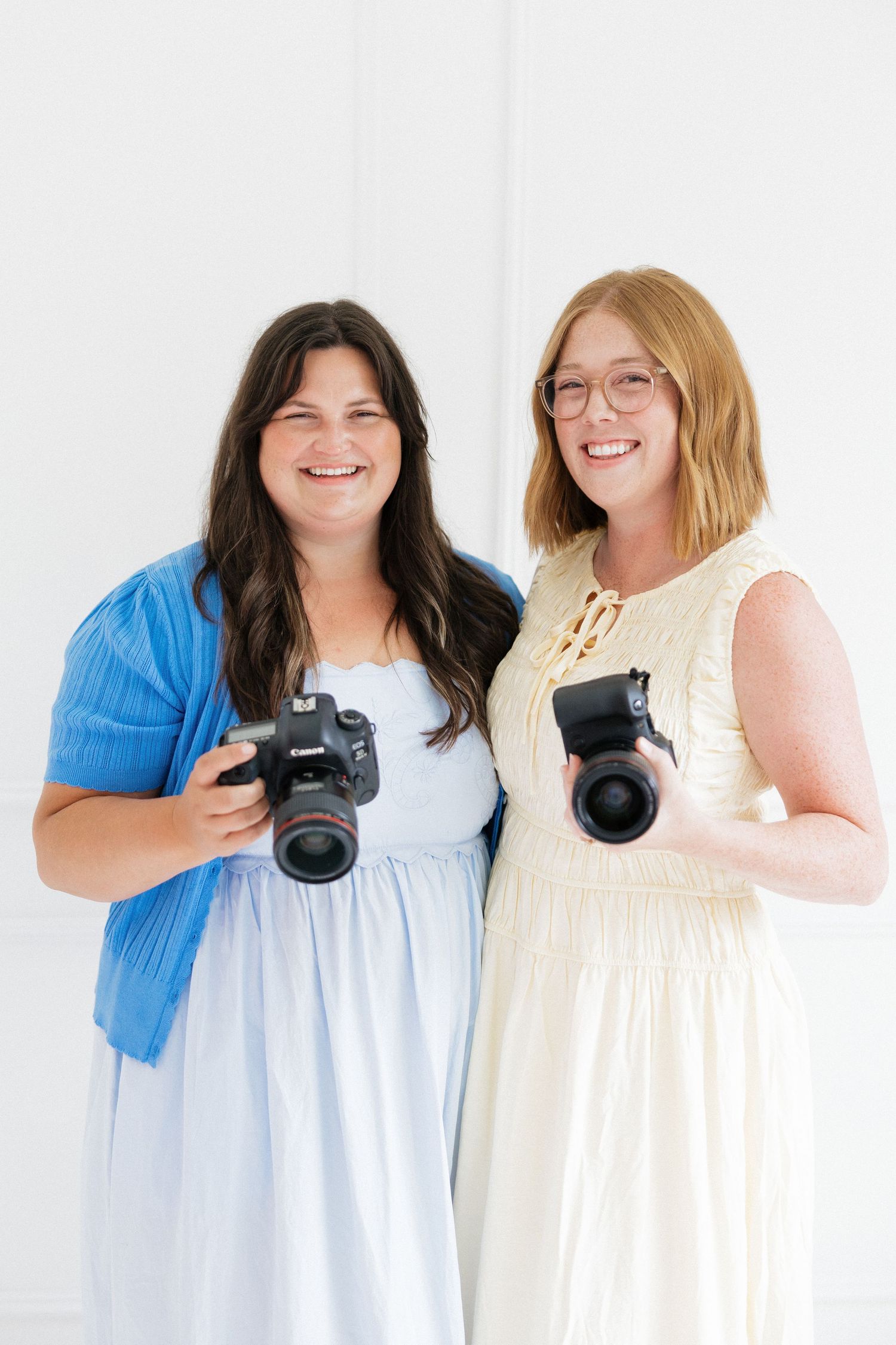 Two cheerful female photographers hold cameras and smile together in a clean white studio environment.