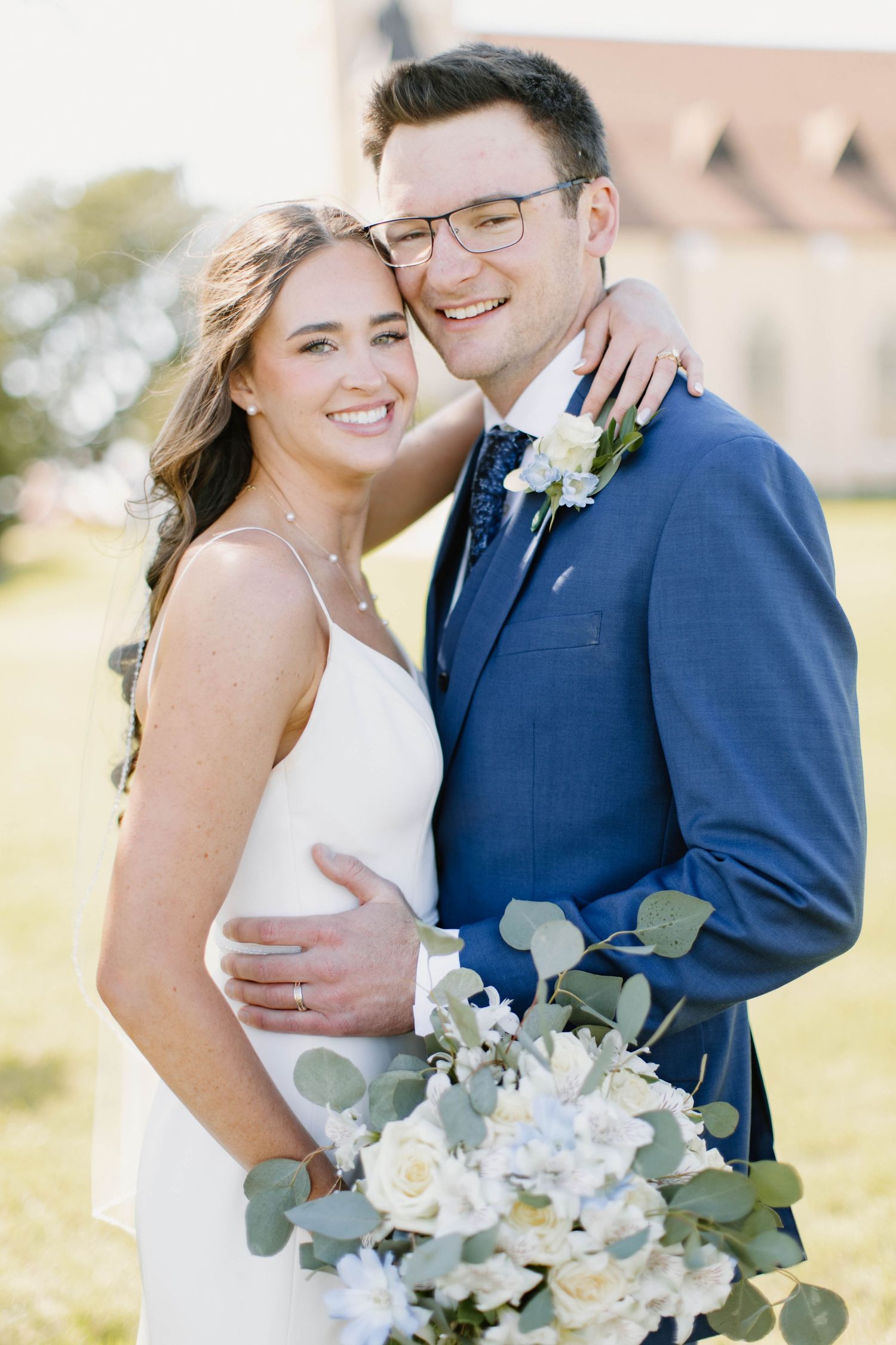 Smiling bride and groom pose outdoors, she holds white and green bouquet while he wears a navy blue suit.
