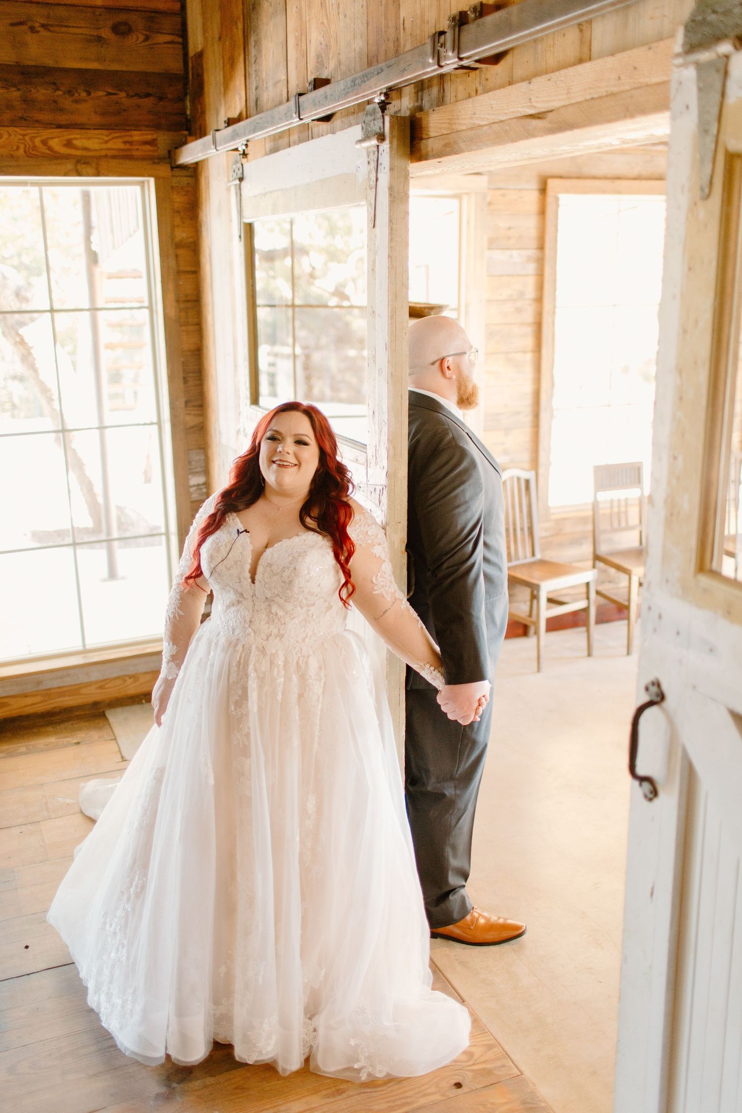 Bride with vibrant red hair in a lace ballgown holds grooms hand during first look in a rustic wooden barn.