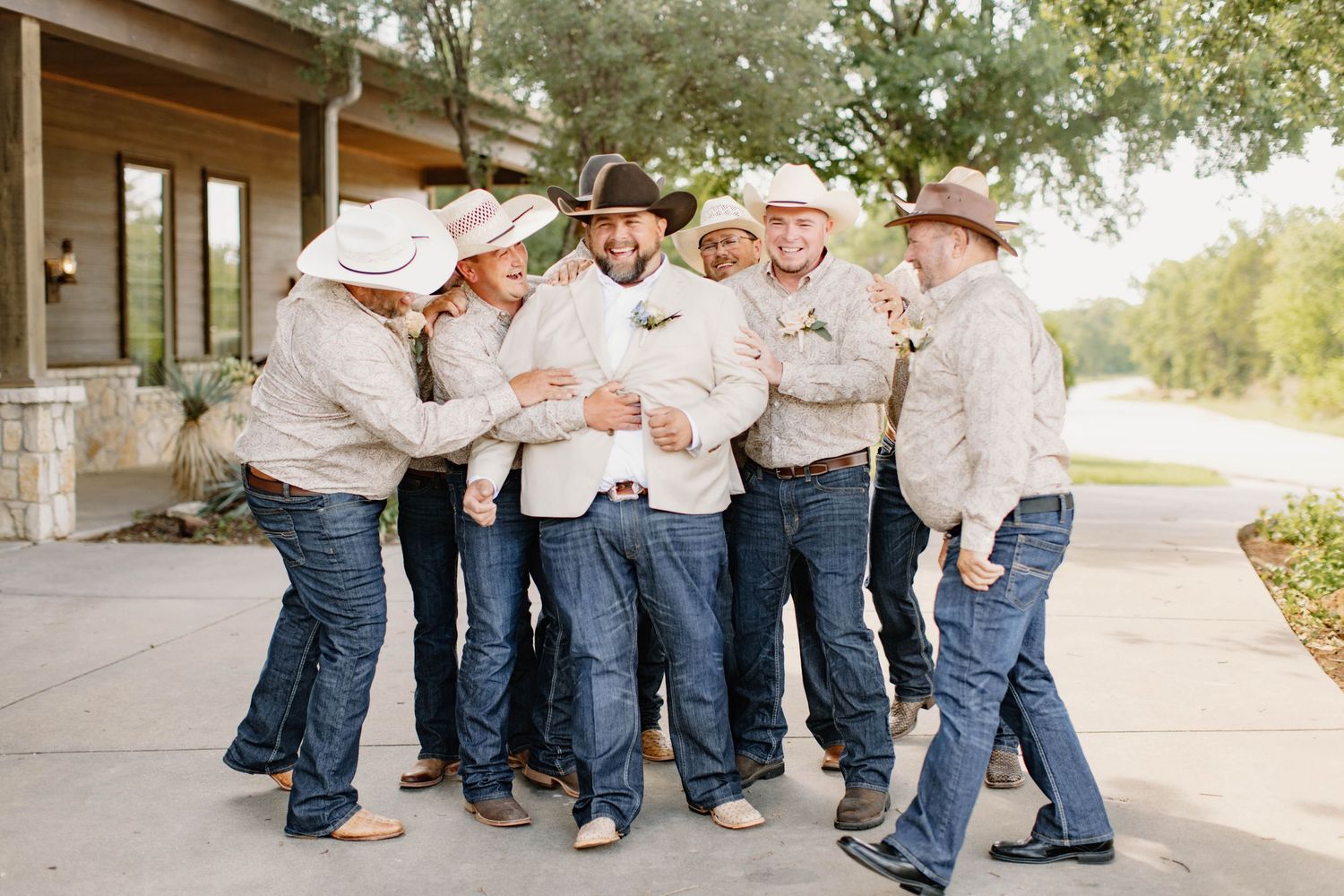 Groom and groomsmen in matching western outfits with cowboy hats and jeans celebrate outdoors at a ranch wedding.