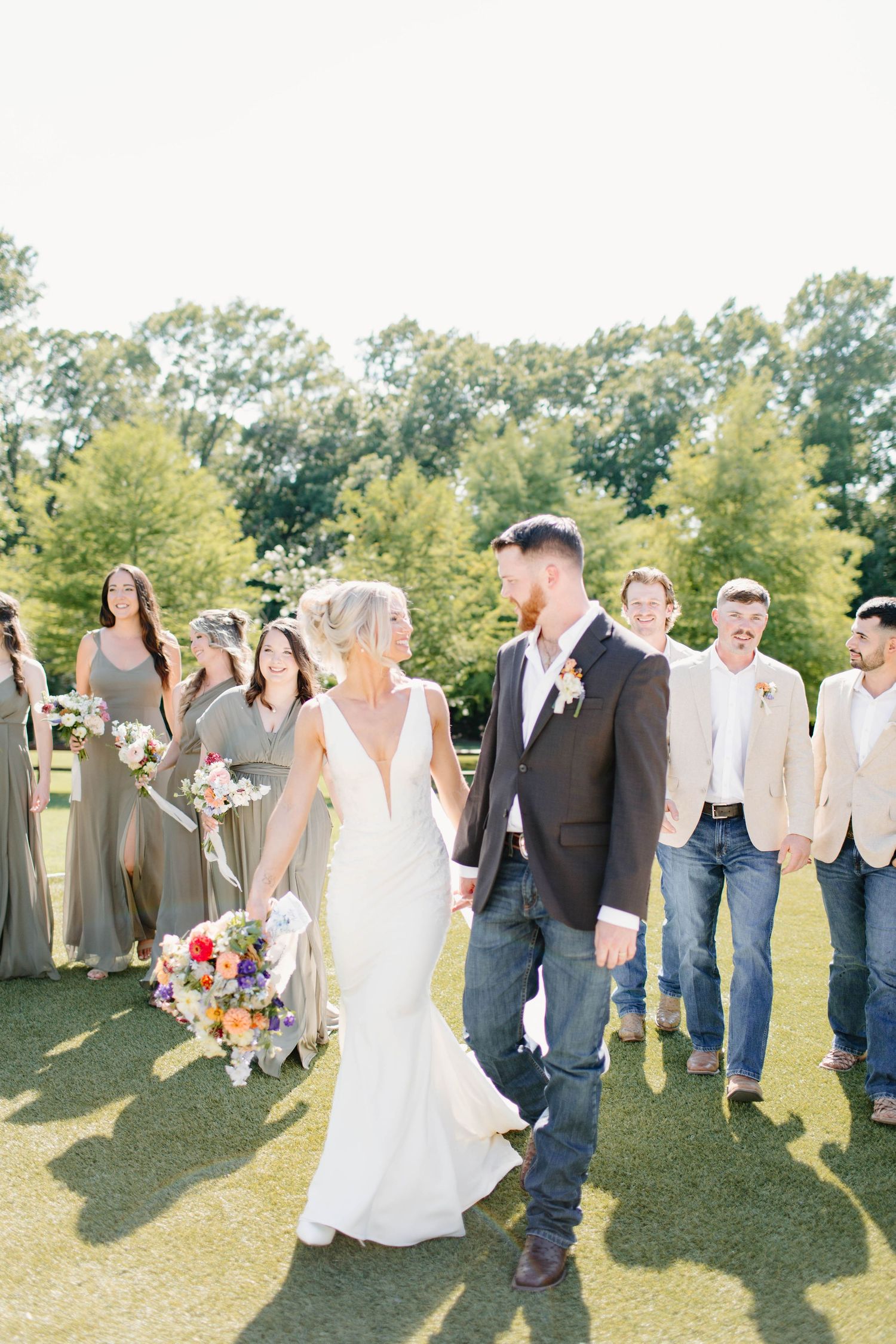 Bride and groom walk hand-in-hand with wedding party in sage dresses and khaki suits on sunny green lawn.