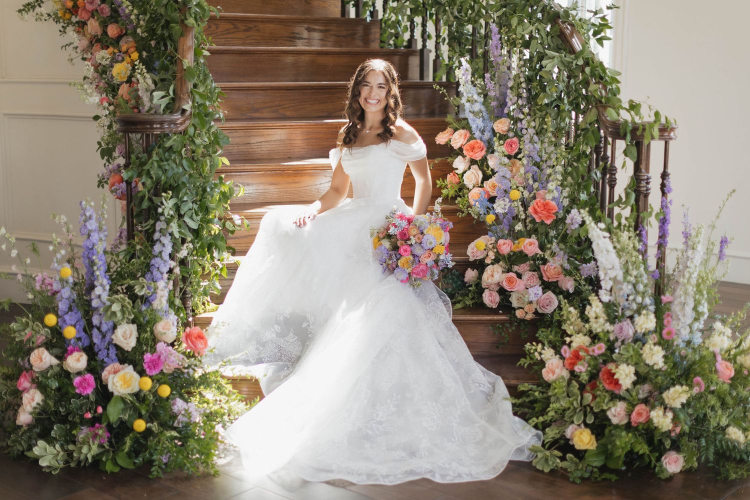 A bride in a white off-shoulder mermaid gown poses on stairs surrounded by colorful floral arrangements.