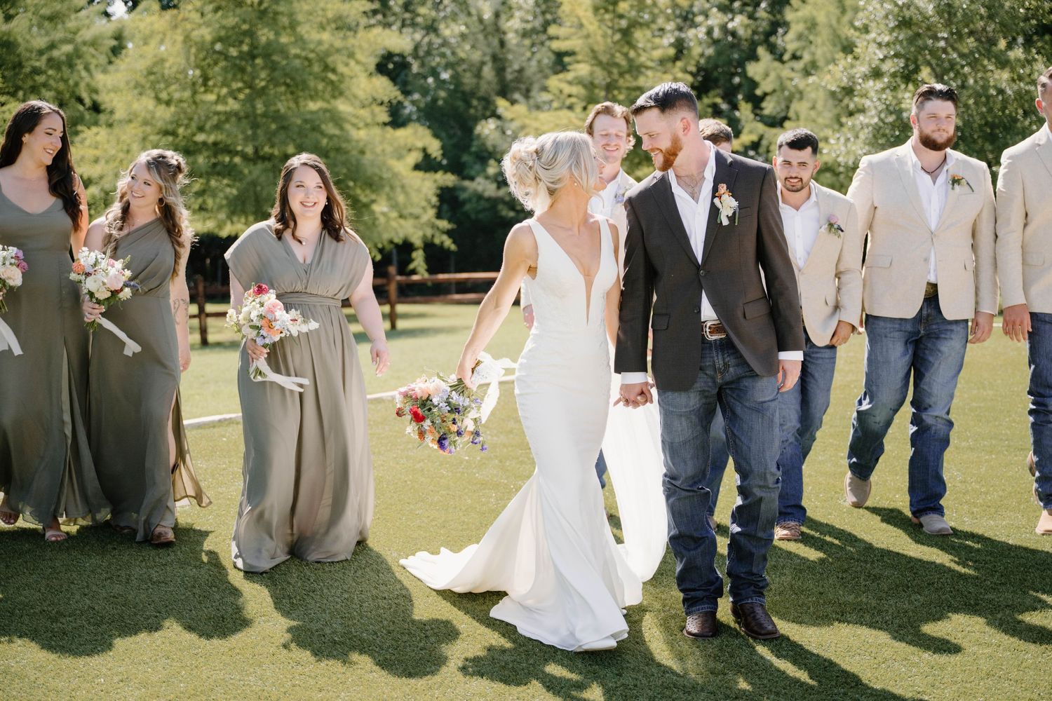 Wedding party walks outdoors on green lawn, bride in white gown with bridesmaids in sage dresses and groomsmen in casual attire.