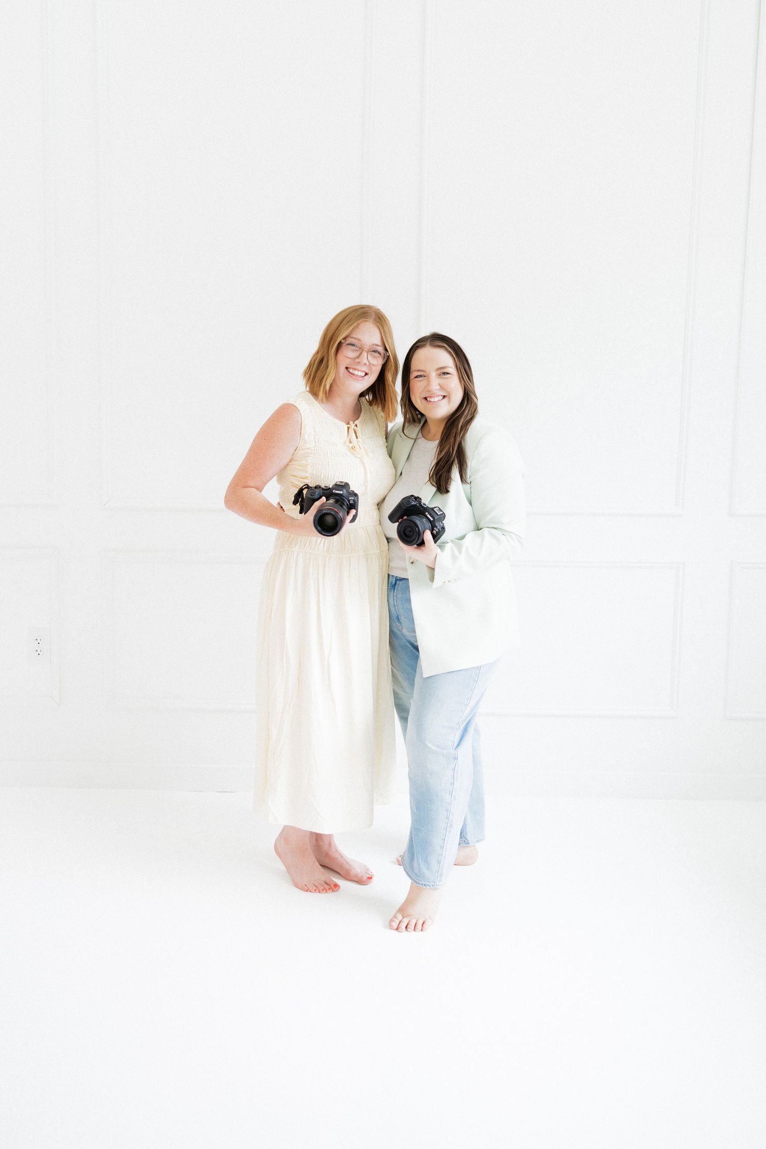 Two barefoot female photographers smile and hold cameras while standing together in an all-white studio space.