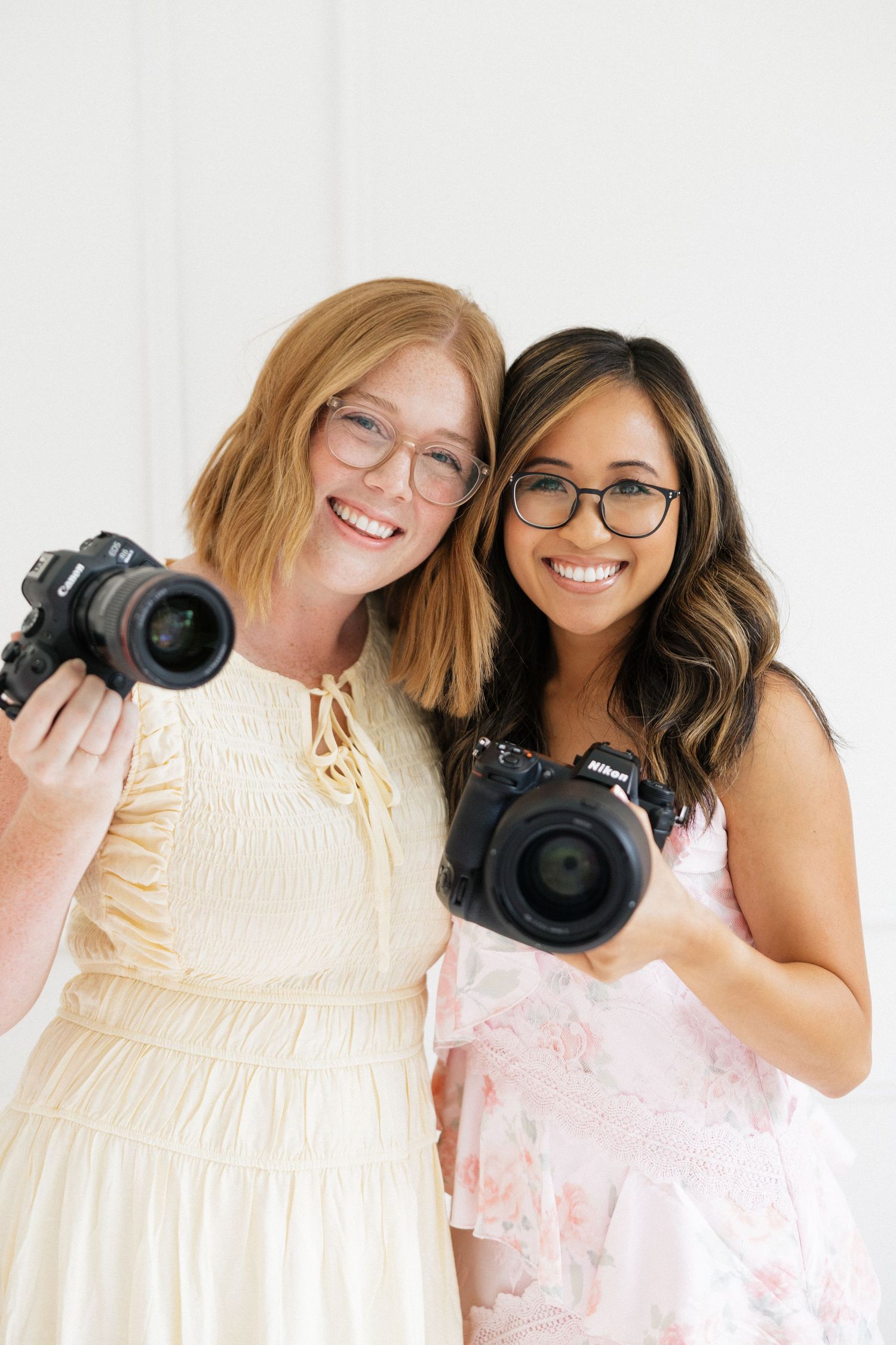 Two smiling female photographers wearing glasses hold Nikon cameras while posing together in a bright studio.