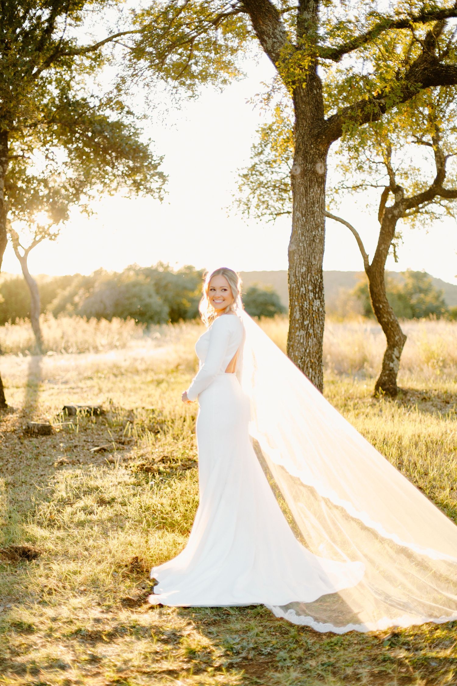 Bride in elegant white gown with flowing veil standing in golden sunlit field at sunset.