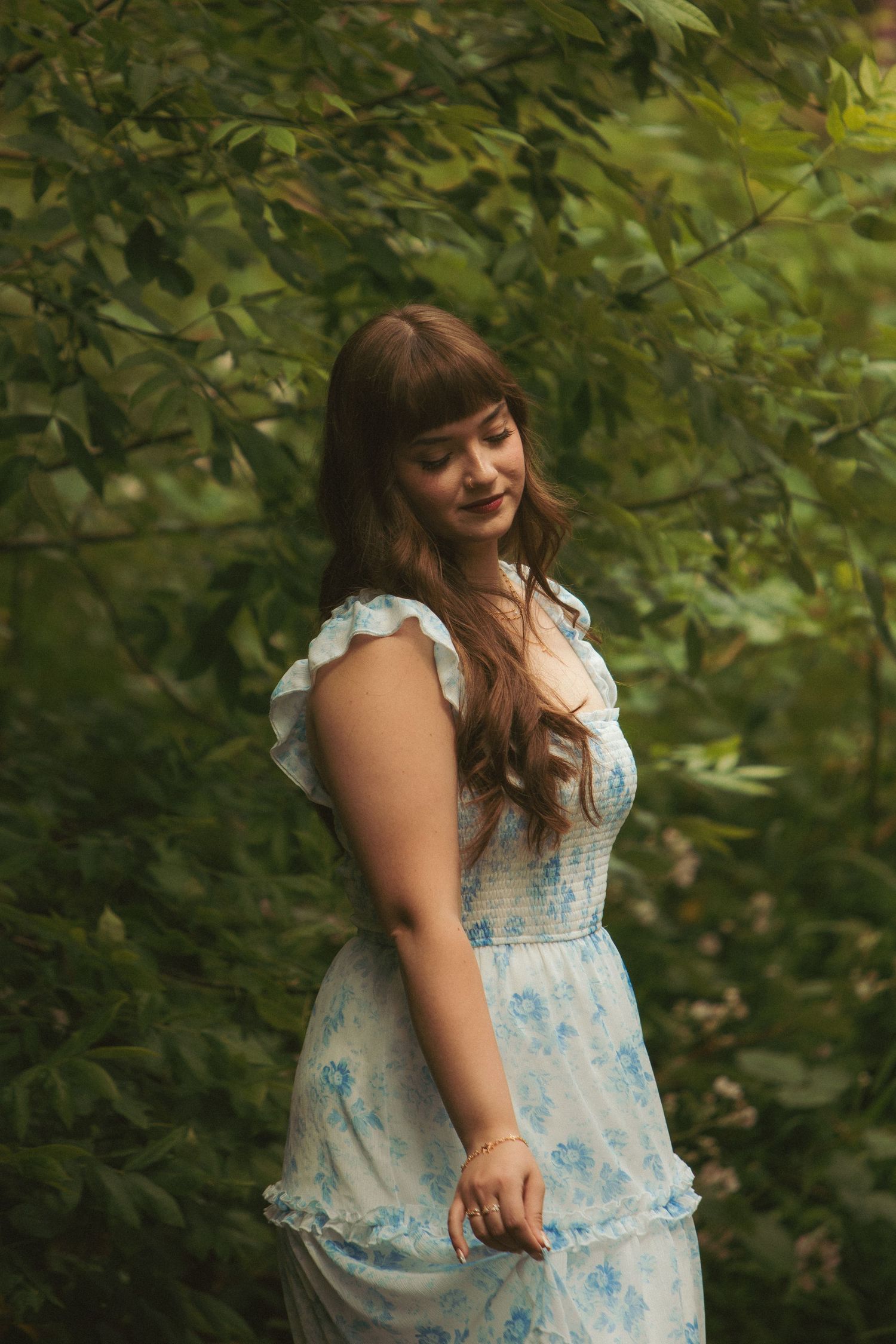 Young woman in a floral blue sundress poses gracefully among lush green foliage in a serene outdoor setting.