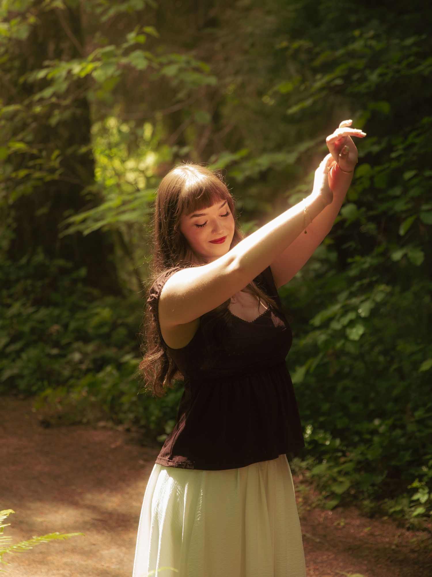 A young woman in a black top and white skirt dances gracefully on a sunlit forest path.