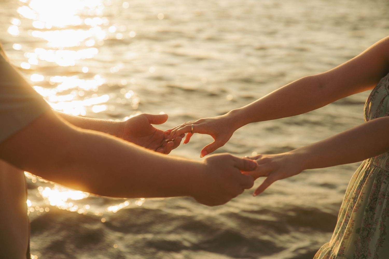 Two people gently reaching toward each other's hands at golden sunset over shimmering ocean water.