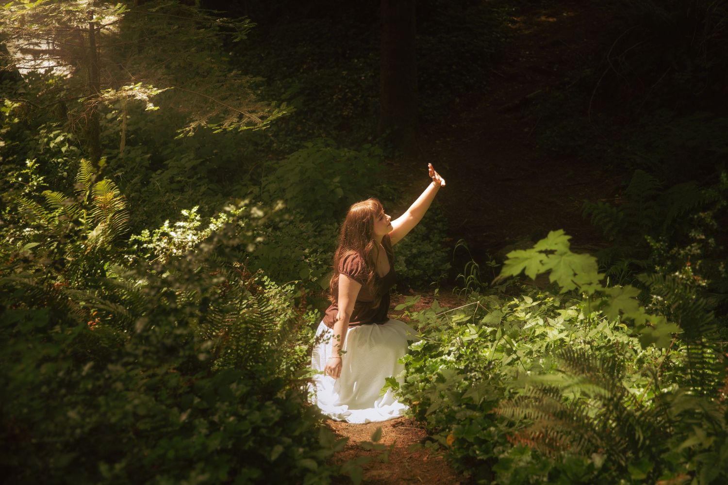 A woman in a white dress reaches up toward sunlight filtering through lush green forest foliage.