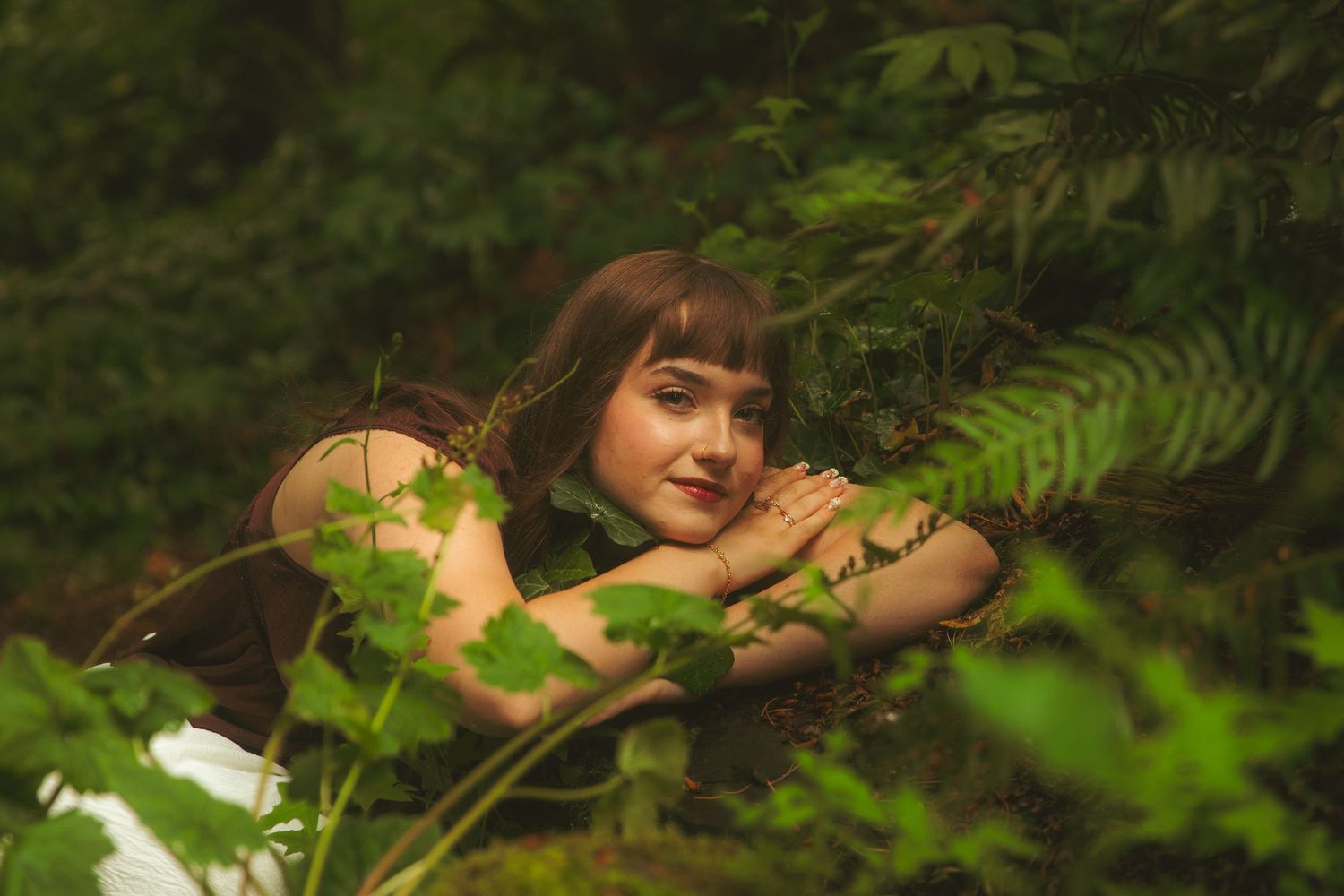 A young woman with bangs rests her chin on her hands, surrounded by lush green ferns and foliage in a forest setting.