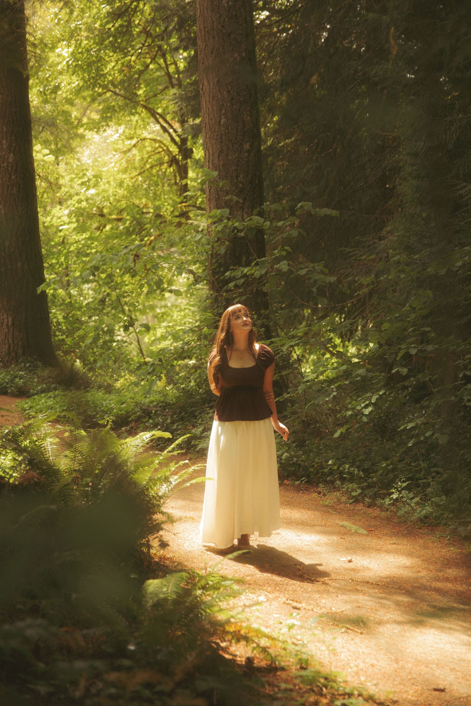 Woman in white skirt and dark top standing on a sunlit forest path surrounded by tall trees and lush green foliage.
