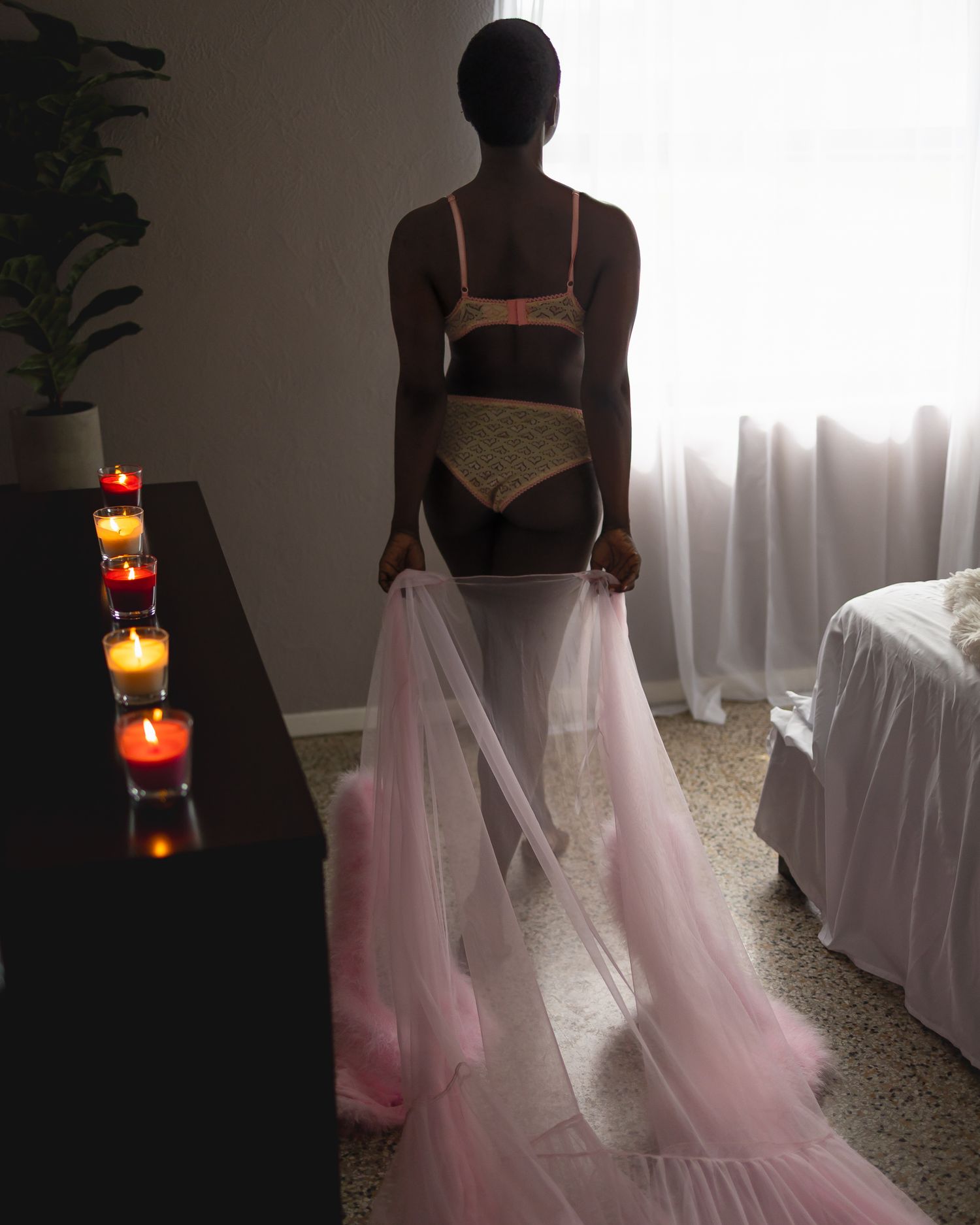 Person in lingerie and pink tulle skirt walking by candlelit table towards a sheer curtained window.