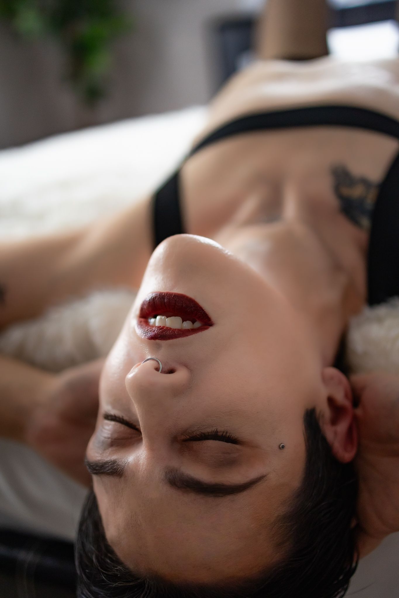 Nonbinary person with red lips and facial piercings lying back with eyes closed, wearing black lingerie with tattoos visible.