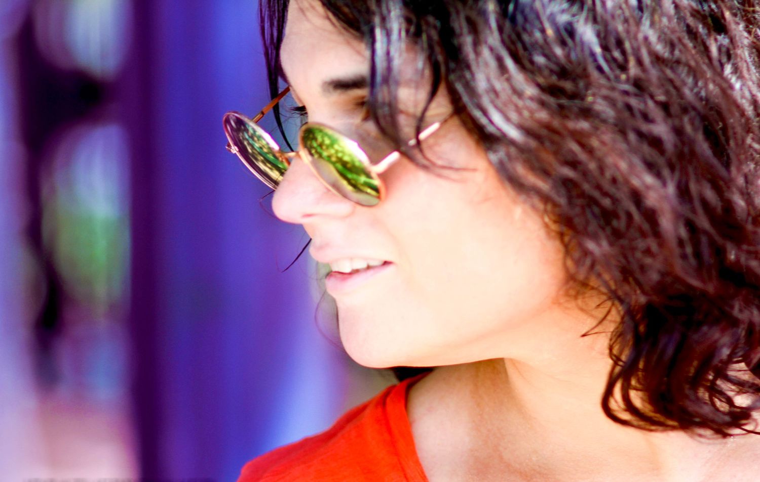 Young woman with curly dark hair smiling in profile, wearing green reflective sunglasses and an orange top.