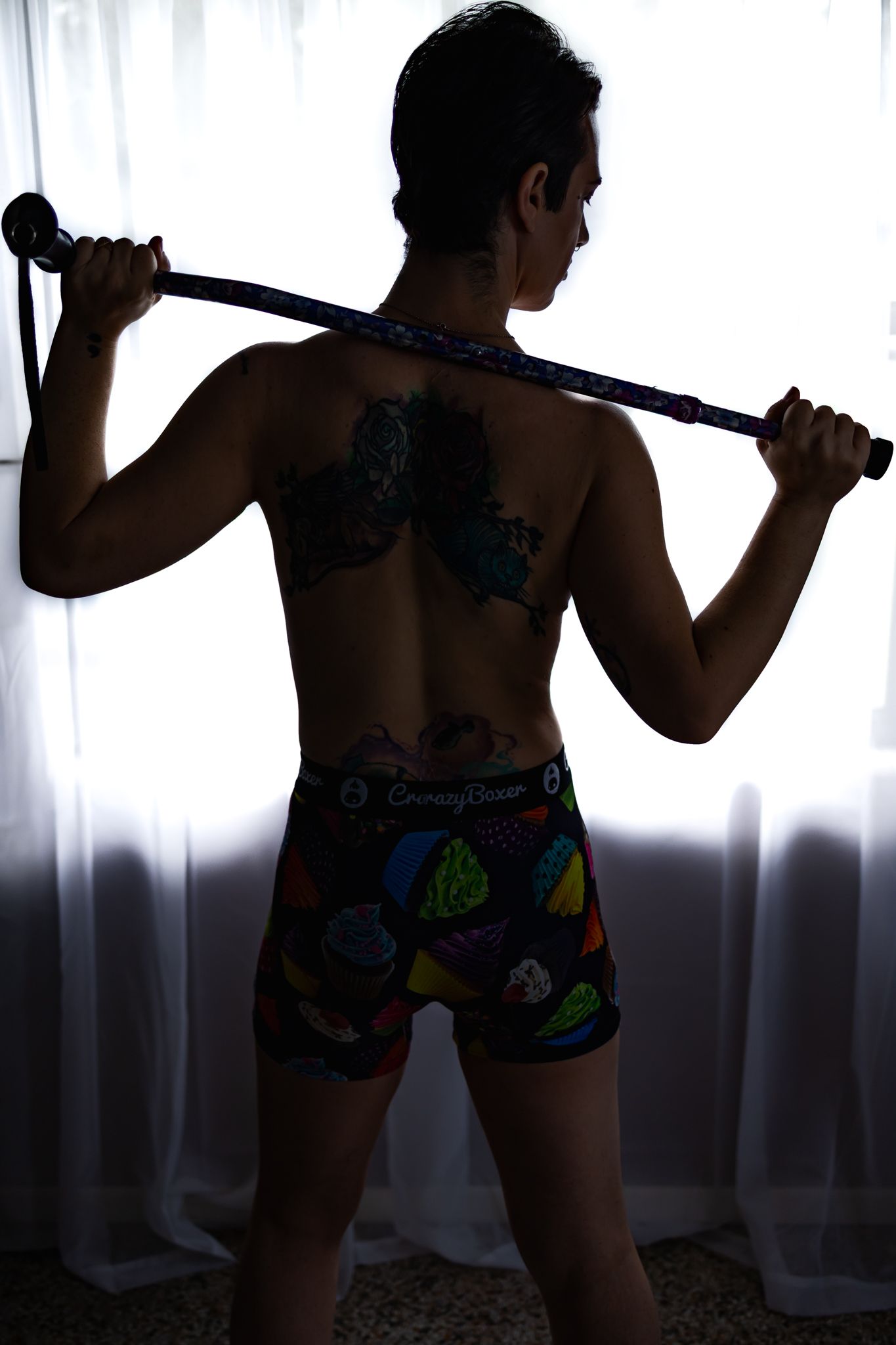 Tattooed nonbinary person holding a walking cane across shoulders, wearing colorful shorts, shown from behind near curtains.