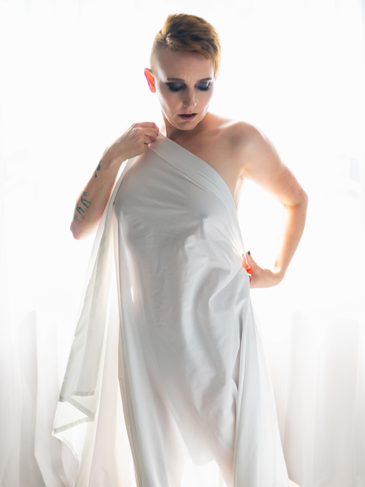Person with short hair and dramatic eye makeup draped in white fabric posing against bright white background.