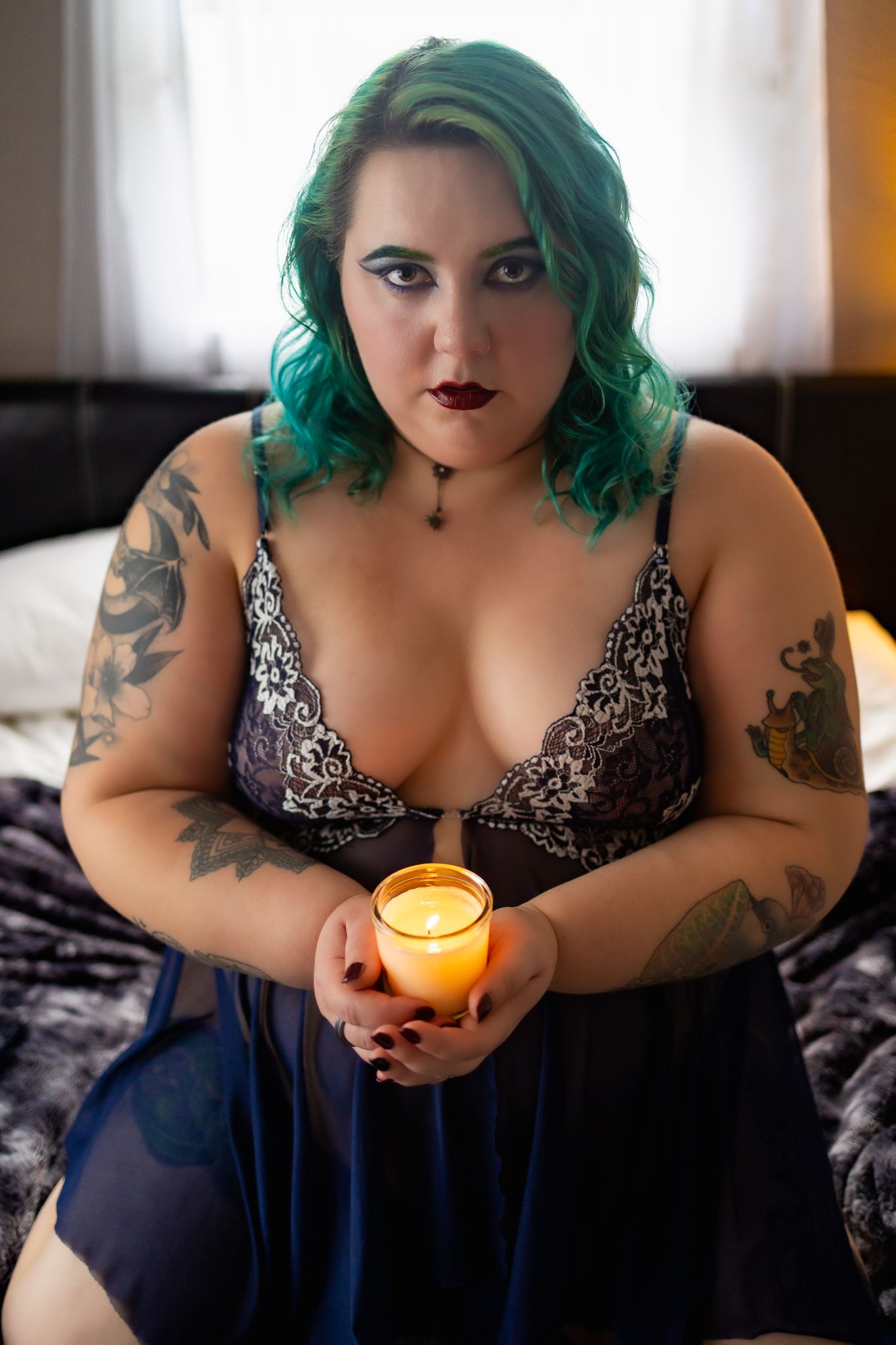 Woman with green hair and tattoos holding a lit candle while wearing lingerie, seated on a bed.